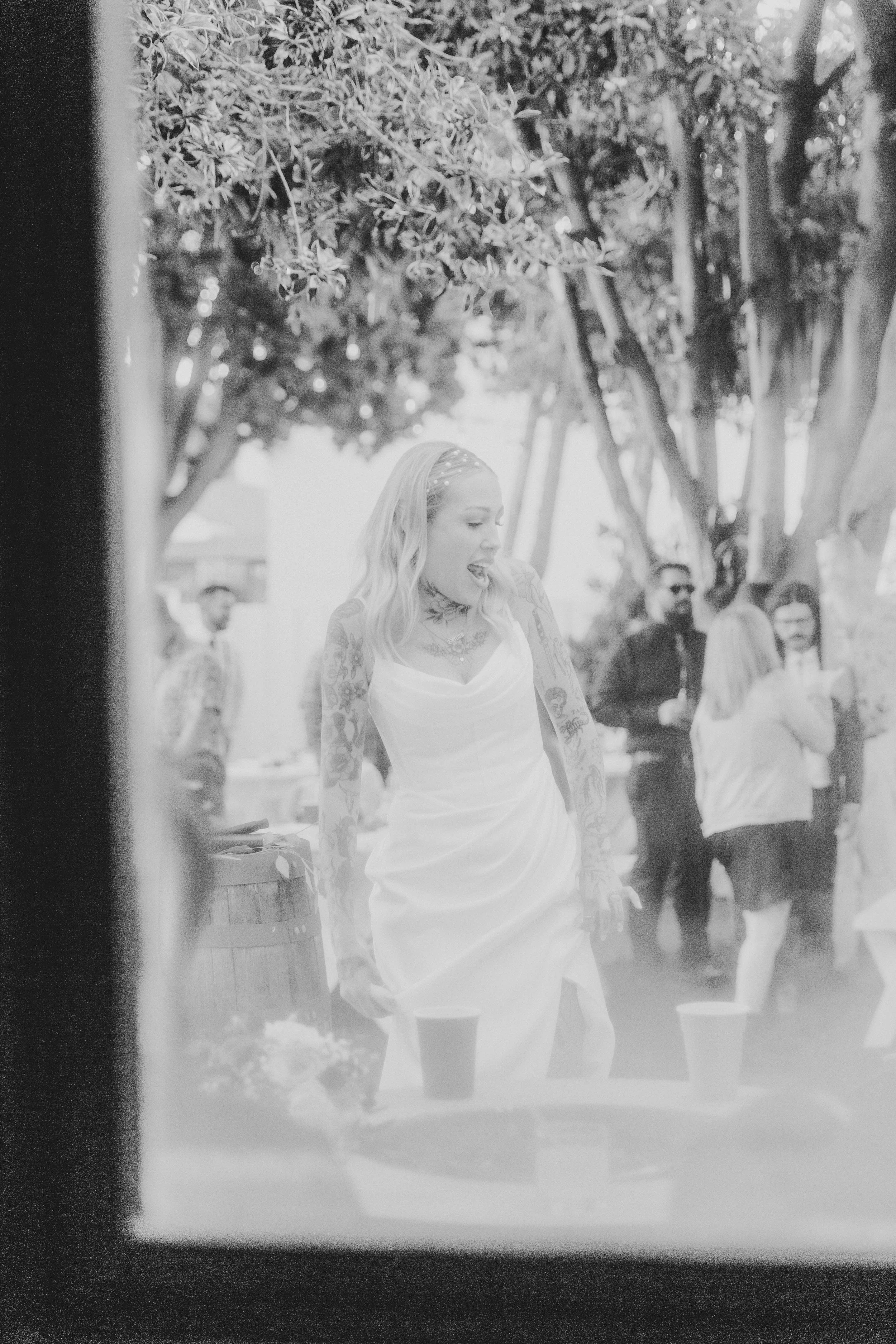 Black and white photo of a bride laughing with guests, seen through a window