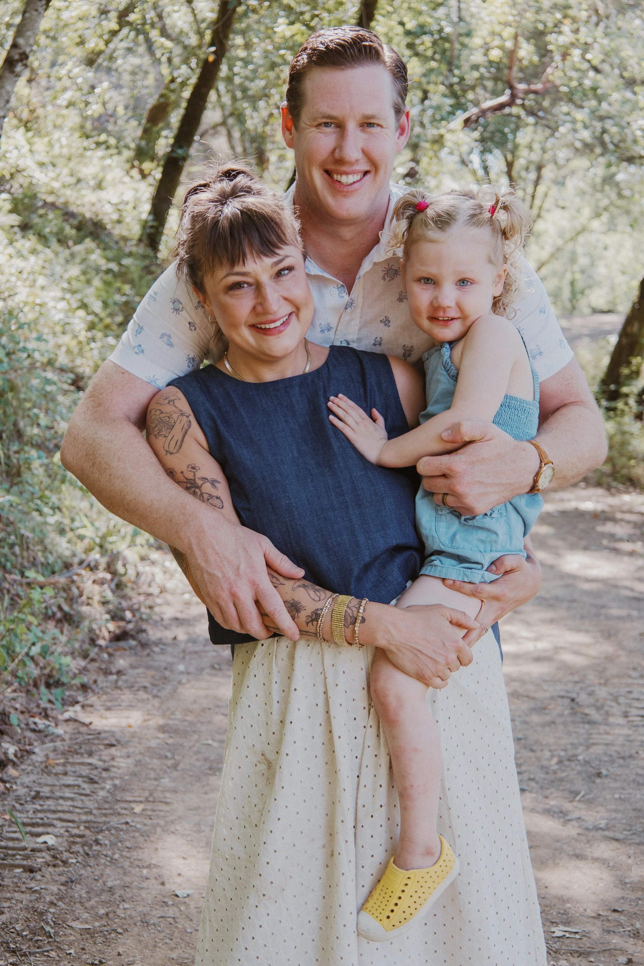 Parents hugging their toddler on a sunny wooded path