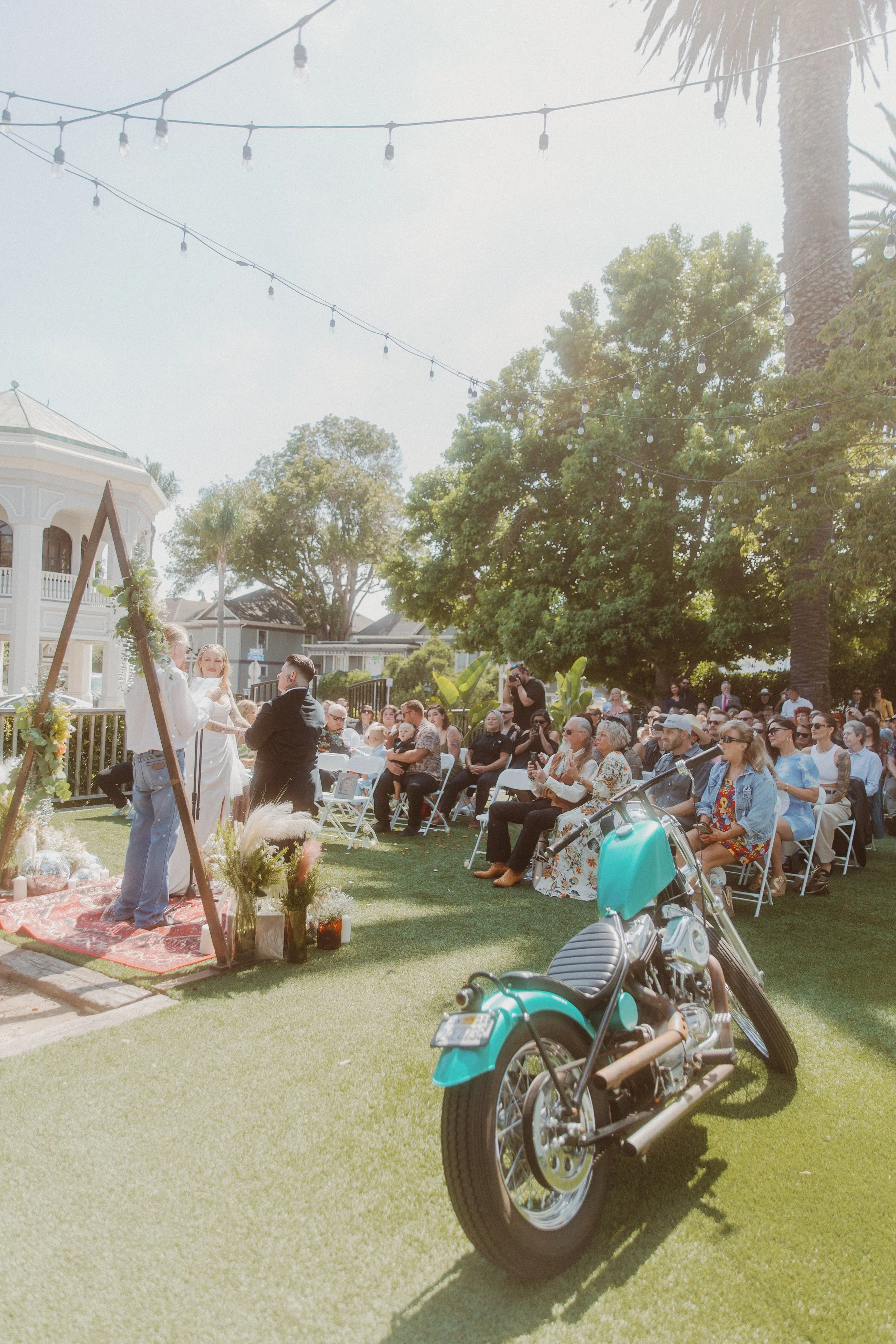 Vintage teal motorcycle parked on the lawn as guests gather for an outdoor wedding ceremony