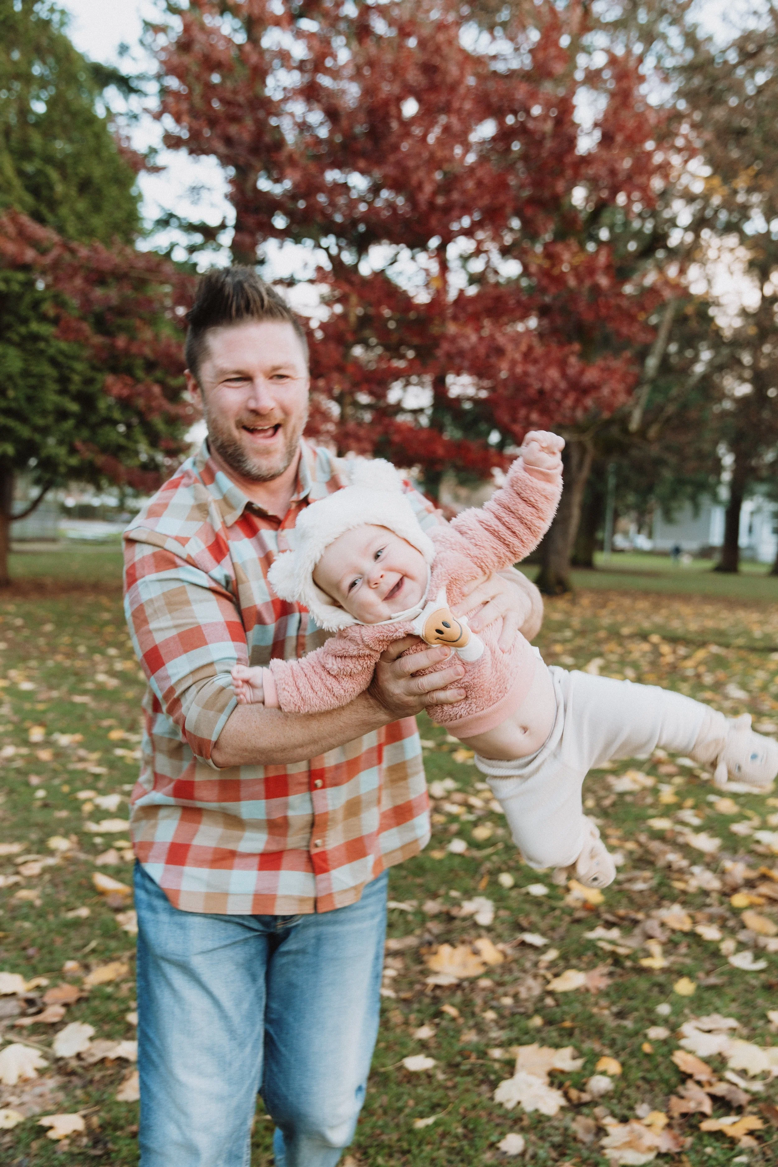 Dad swinging his laughing baby through the air in a park with red fall trees