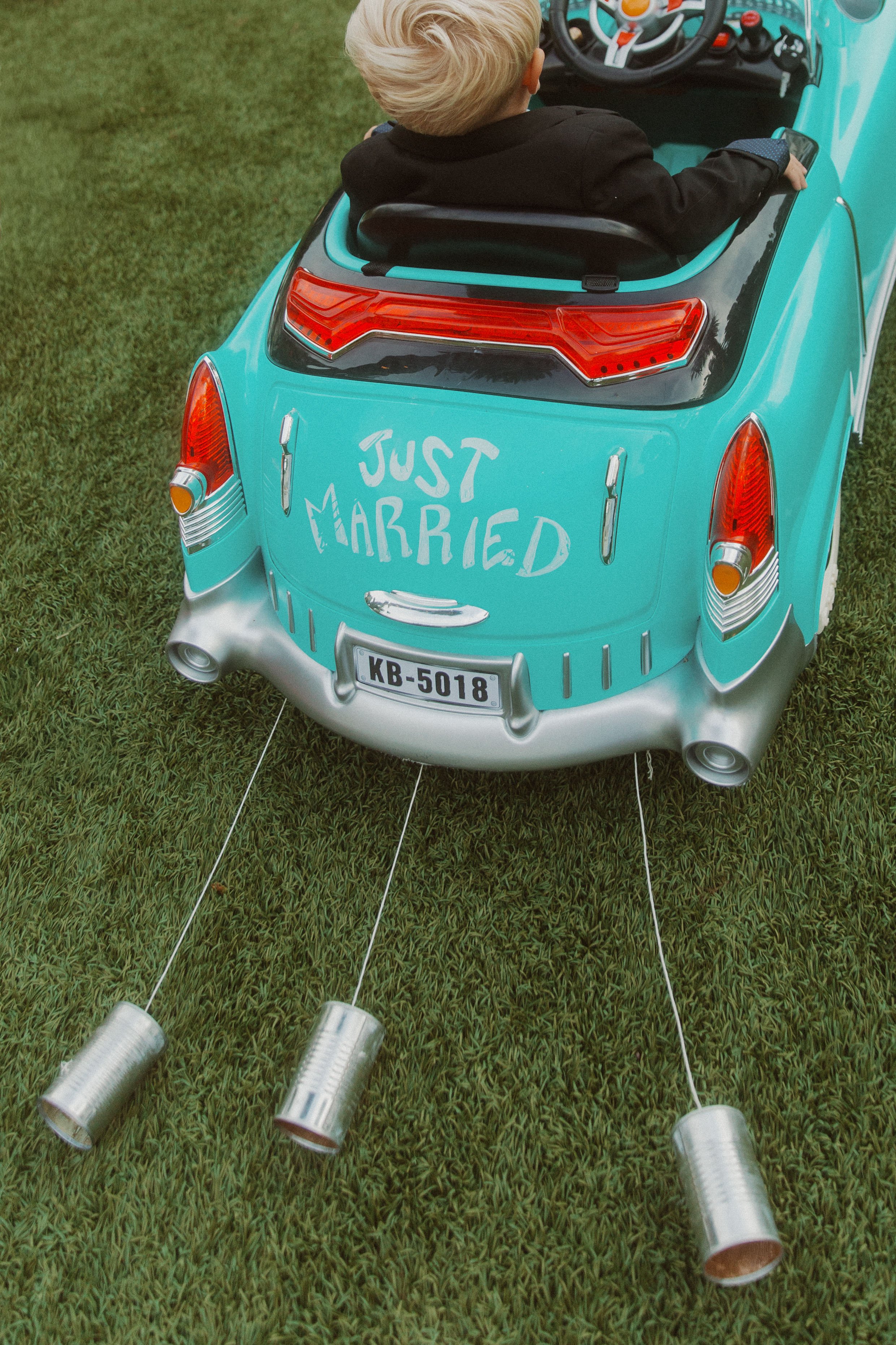 Toddler riding a teal toy car with a Just Married sign and tin cans trailing behind