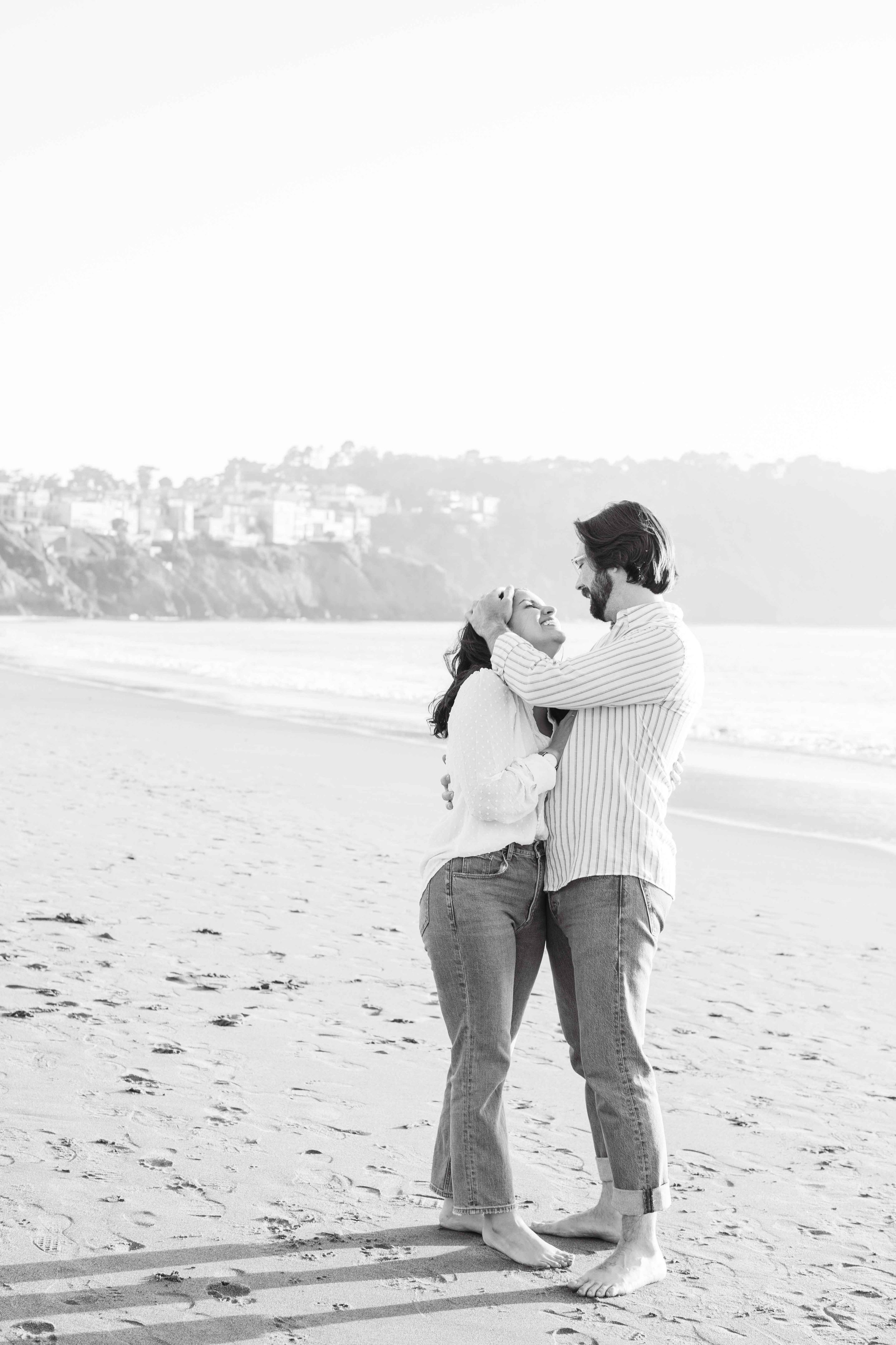 Couple hugging on the beach with the coastline behind them