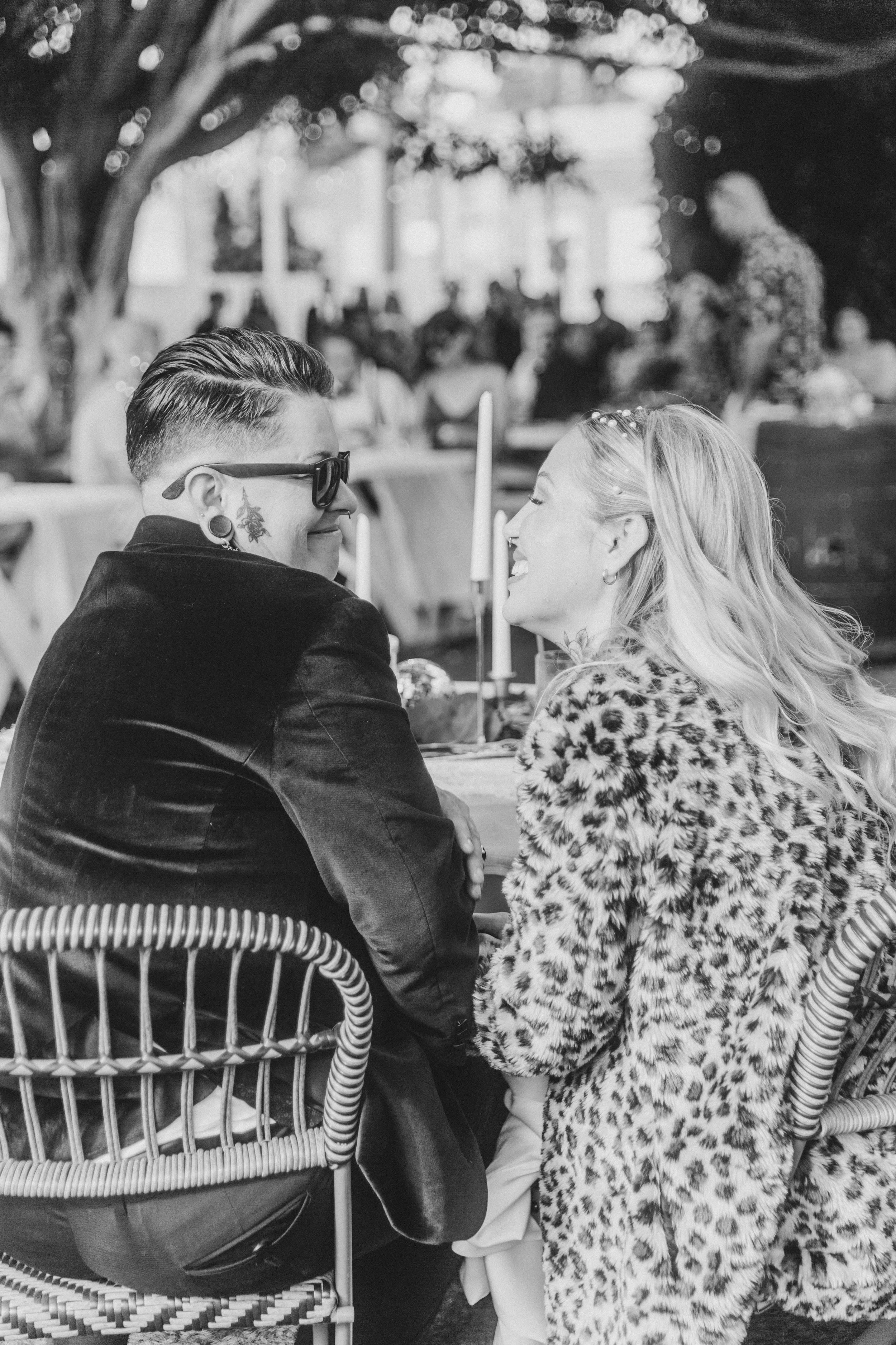 Black and white photo of the newlyweds seated together at their reception, seen from behind