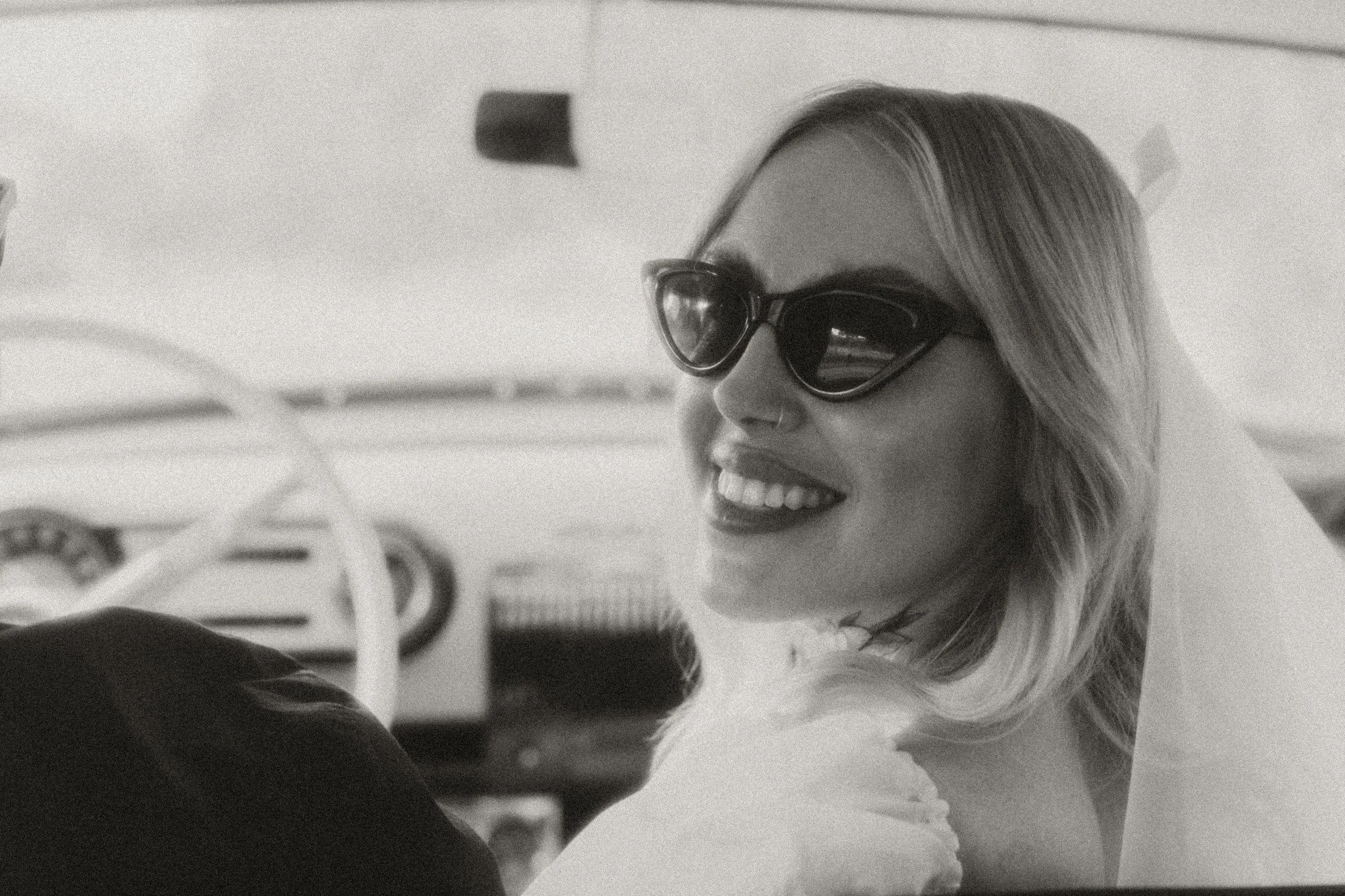 Black and white photo of a bride in sunglasses smiling from a vintage car