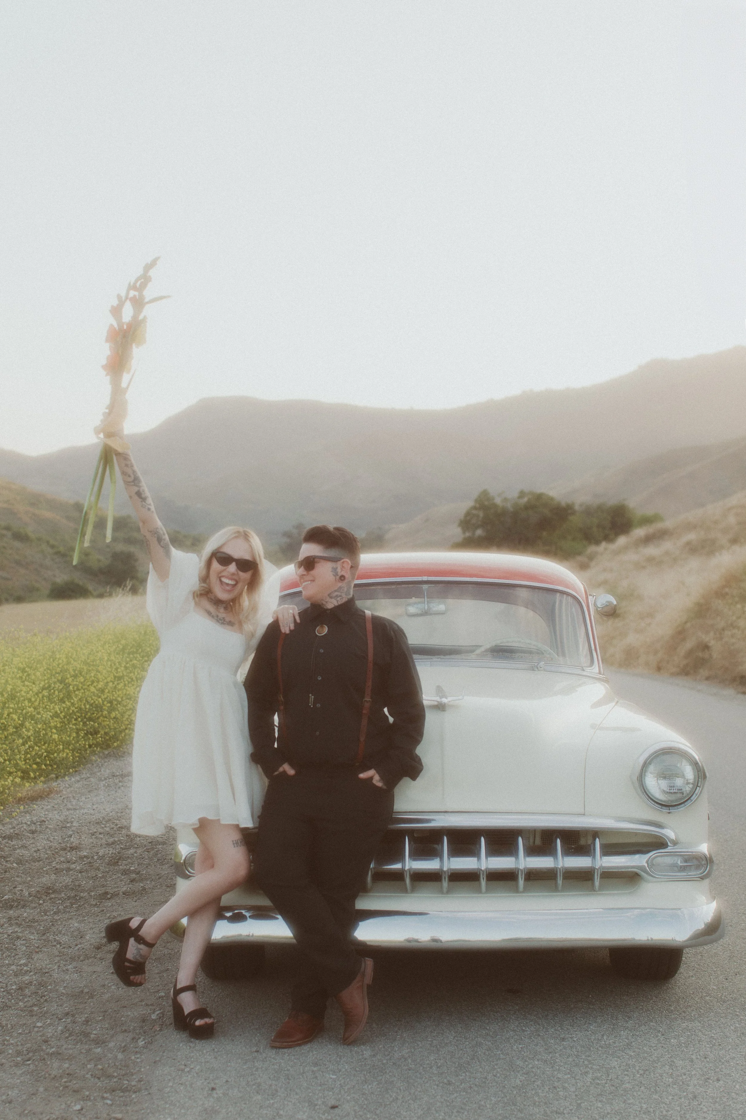 Newlyweds celebrating beside a vintage car on a hillside road