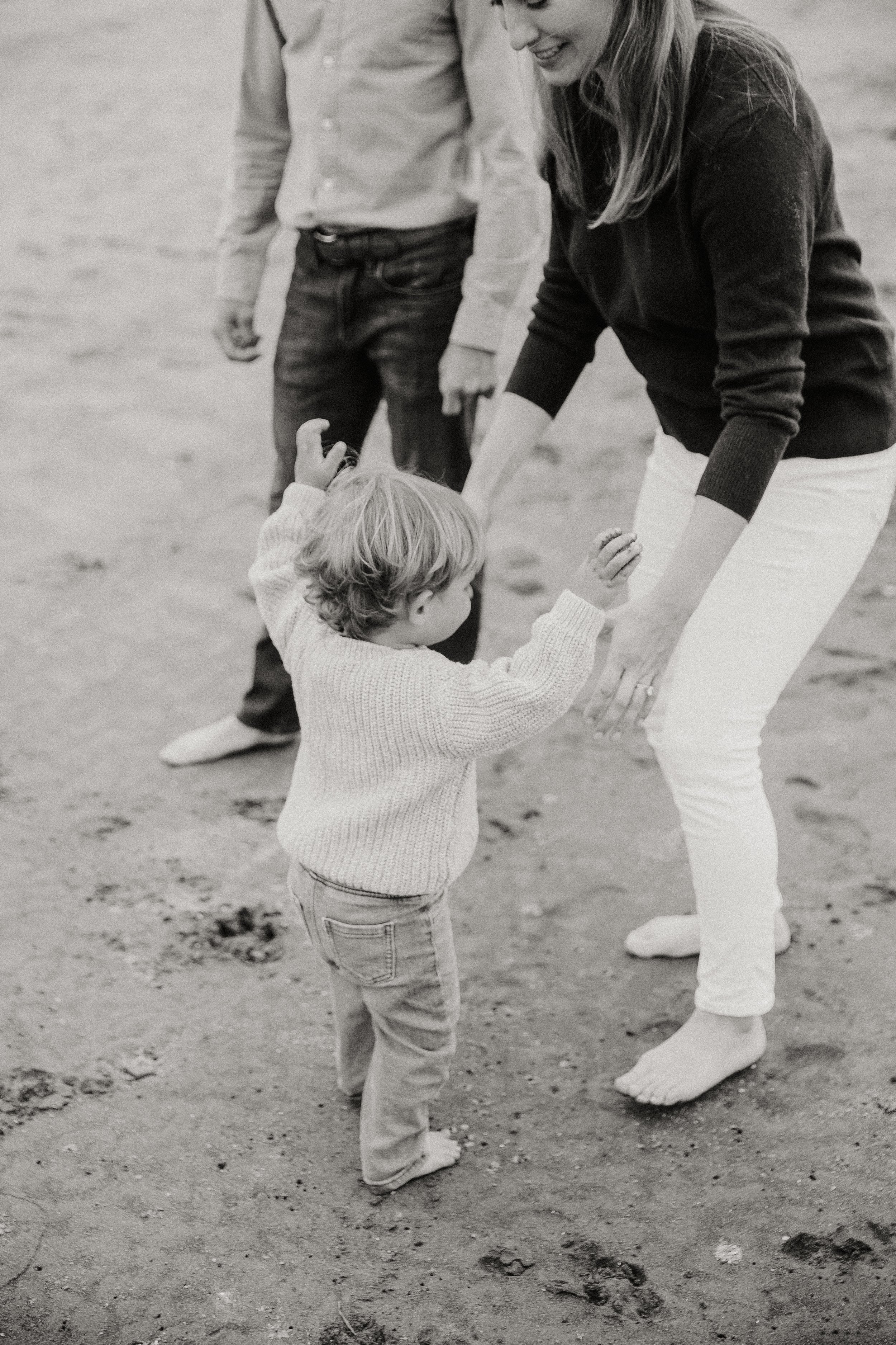 Black and white photo of a toddler walking with parents on the beach