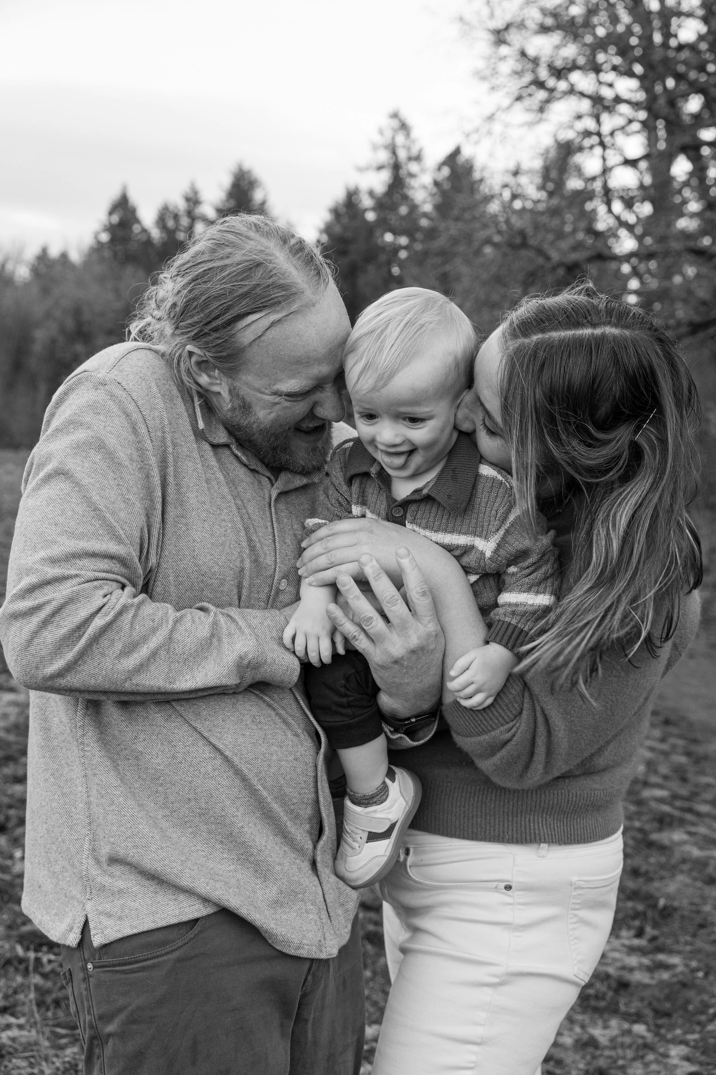 Black and white photo of parents kissing their toddler in a field by the woods