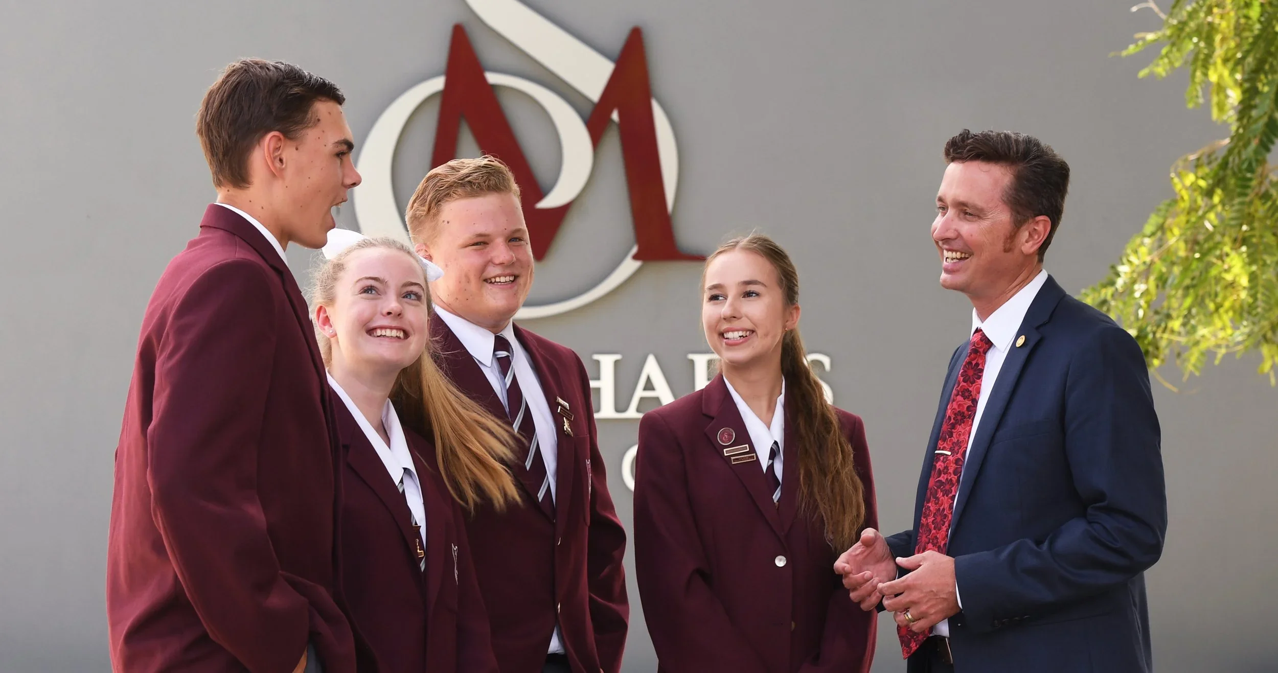 A group of four students in maroon blazers and a man in a navy suit, standing outdoors in front of a university sign, engaging in conversation and smiling. School and Education Marketing Photography