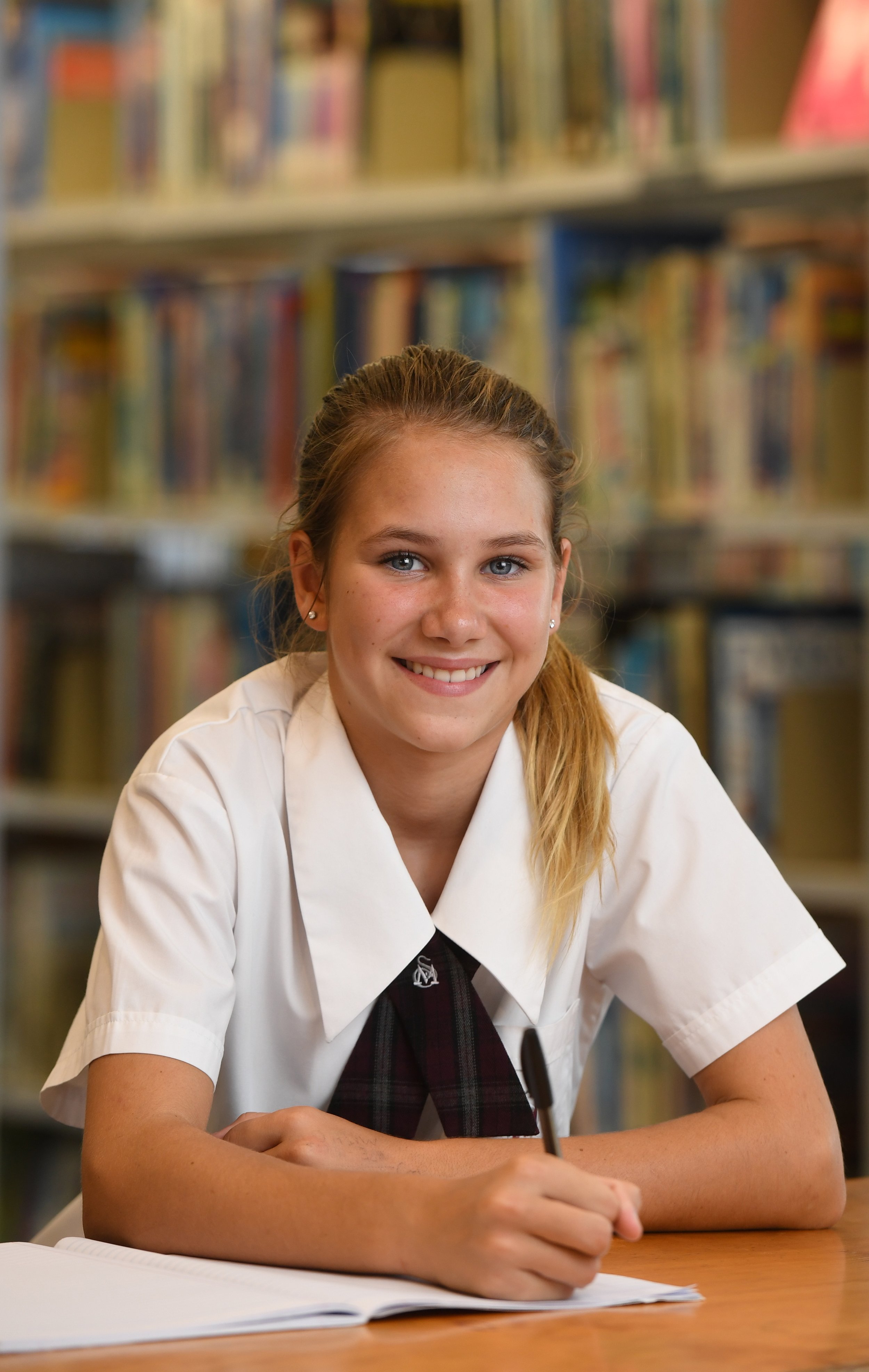 A young girl with long, blonde hair in a ponytail, sitting at a table with an open notebook, smiling and holding a pen, in a school library with shelves of books in the background.