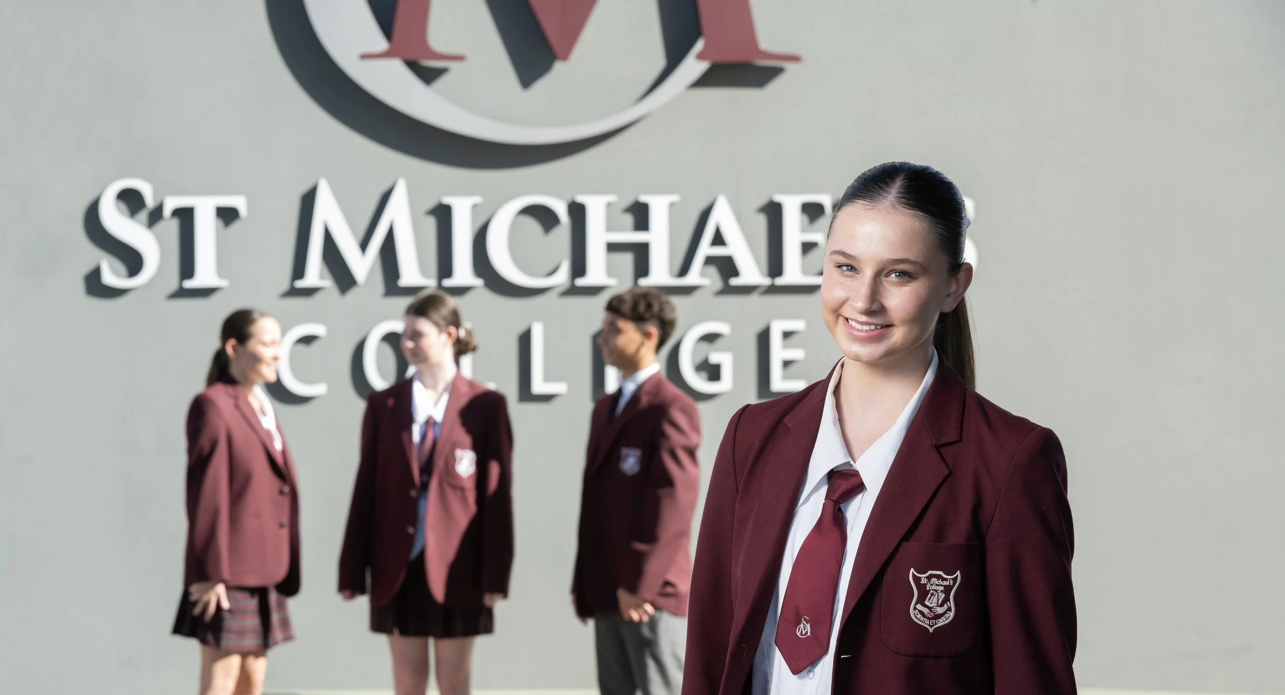 Young woman in a maroon blazer and tie standing outside St. Michael's College with a smiling face, four students in similar uniforms engaging in conversations in the background