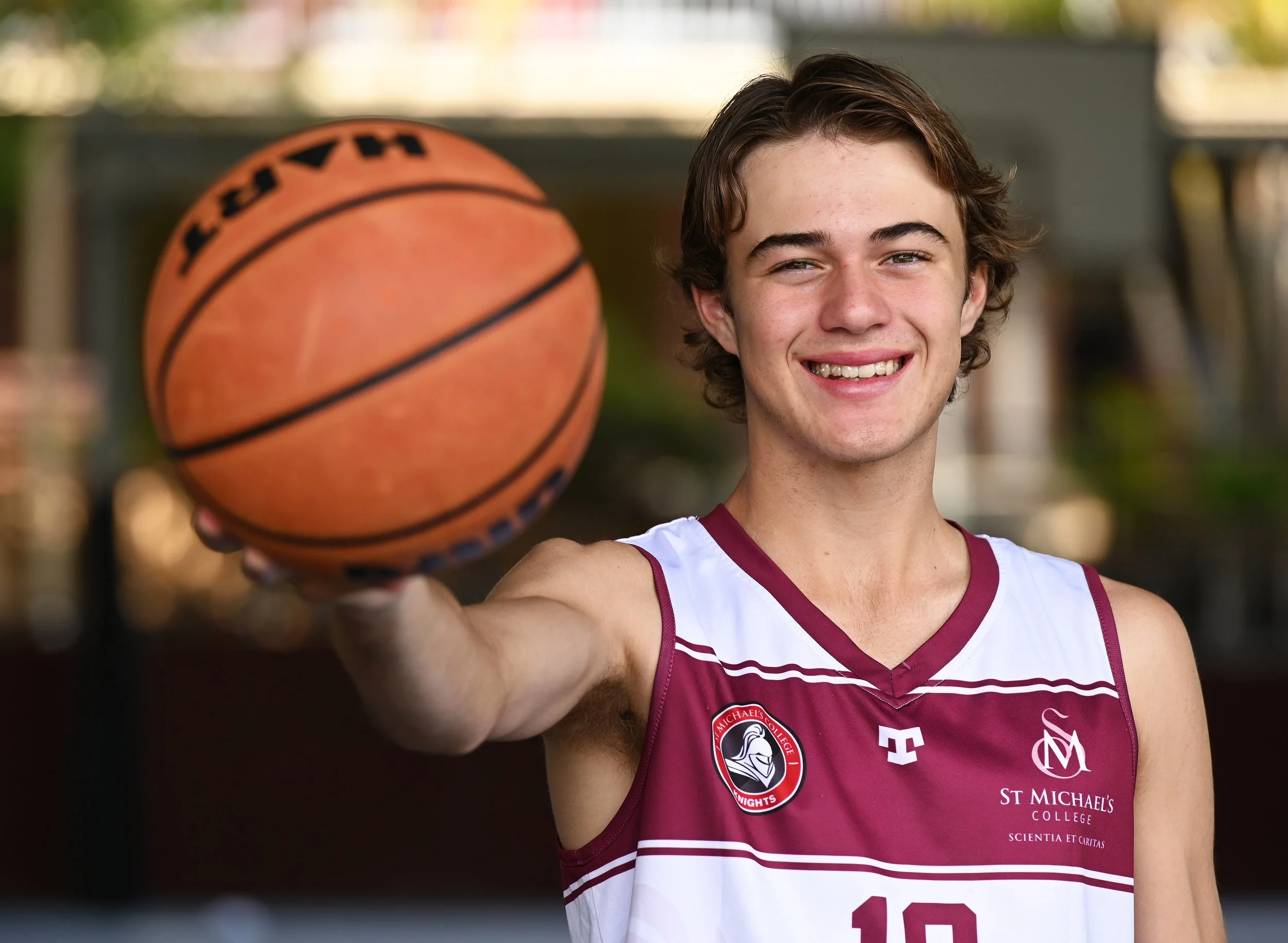 A young male basketball player with light skin and curly brown hair, smiling and holding a basketball toward the camera. He is wearing a maroon and white basketball jersey from St. Michael's College.