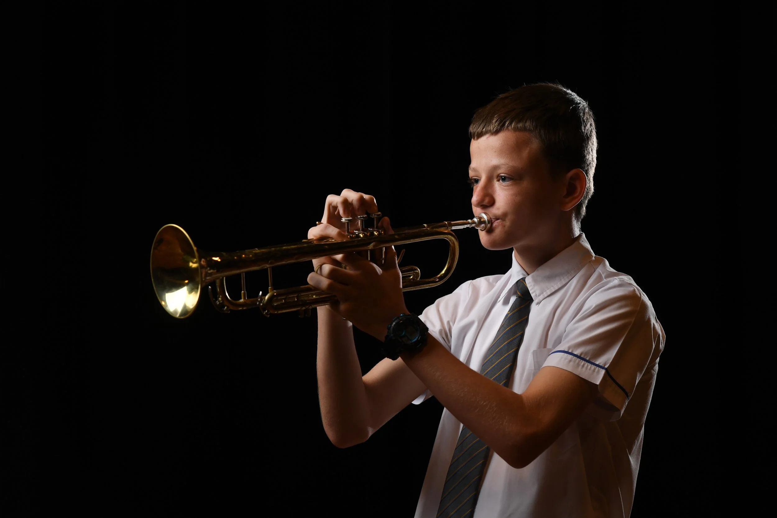 A young boy wearing a white shirt, striped tie, and black watch playing a trumpet against a black background.