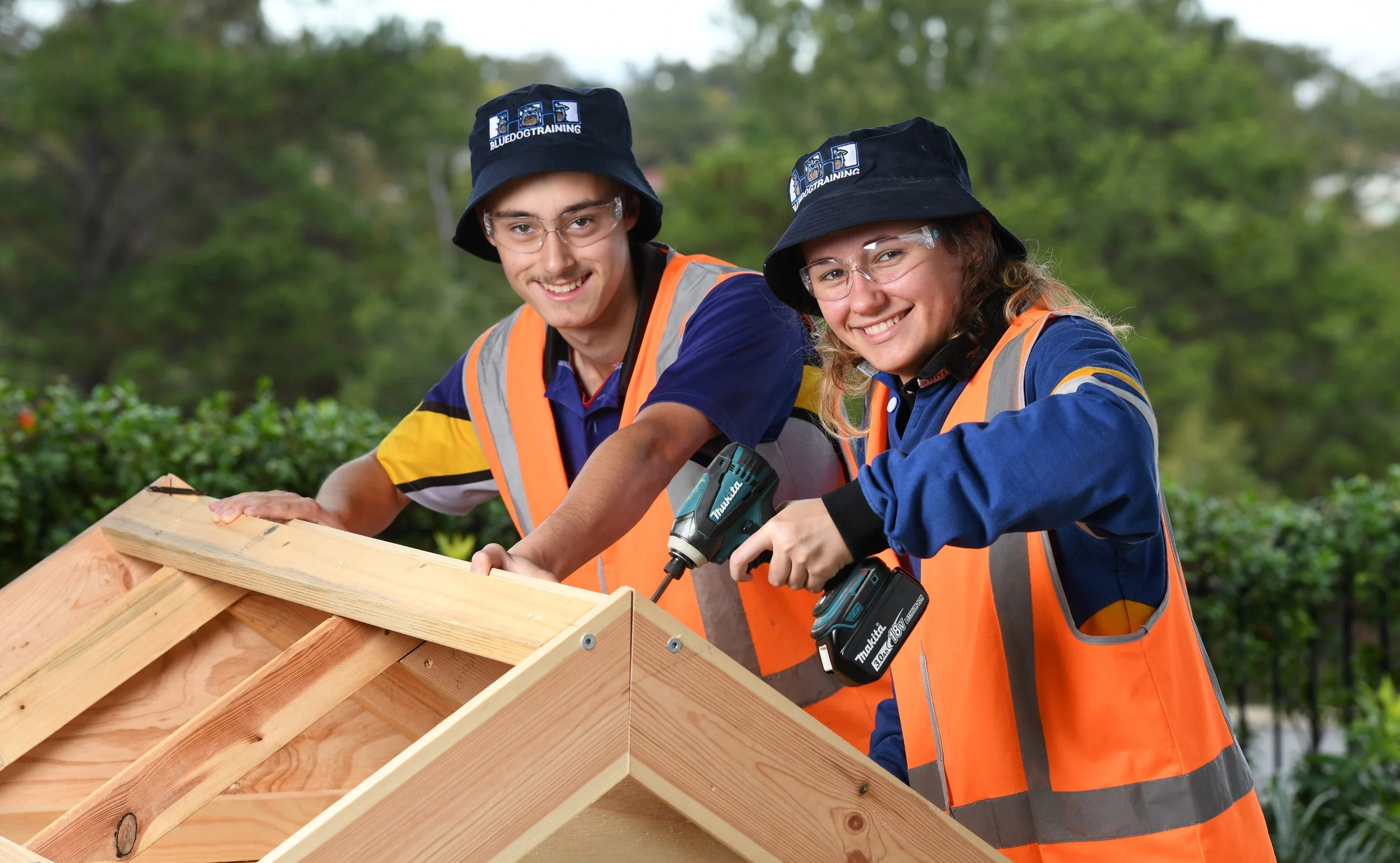 Two young craftsmen, a boy and a girl, working together on a wooden roof structure outdoors. They are wearing safety vests and hats. School and Education Marketing Photography.