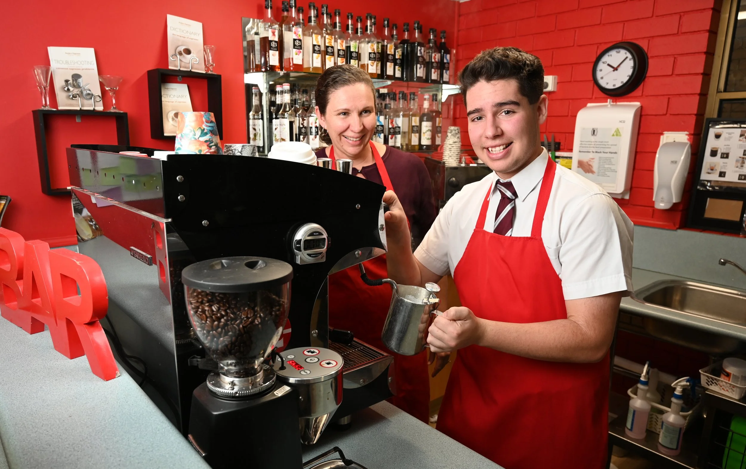 A young man in a white shirt, red apron, and tie making coffee behind the counter of a café, with a woman in a red apron smiling in the background, red brick walls, coffee bottles, and equipment visible.