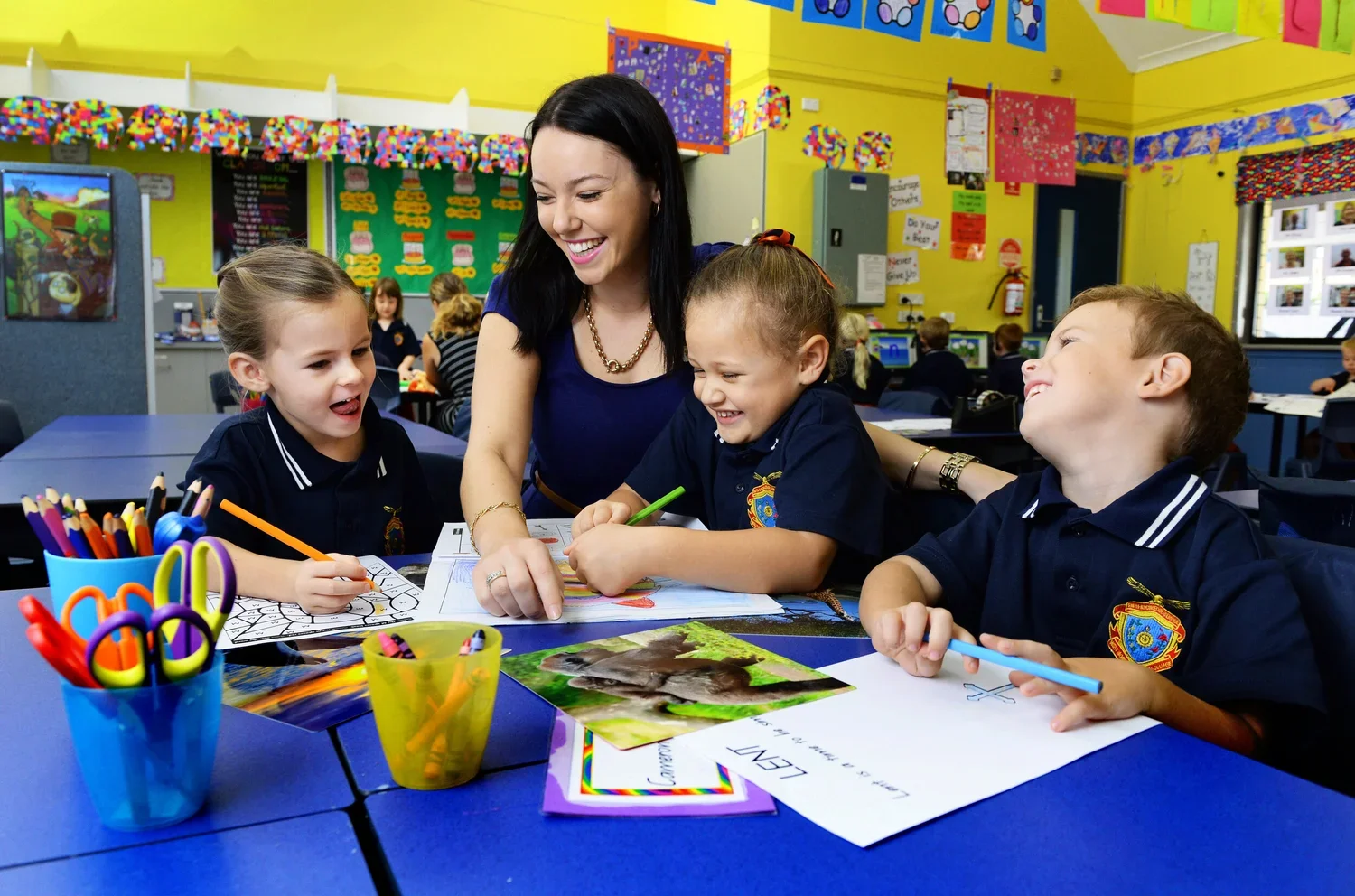 A teacher interacting with three young students in a colorful classroom. The children are smiling and drawing with colored pencils at a blue table, with various art supplies around them. School and Education Marketing Photography