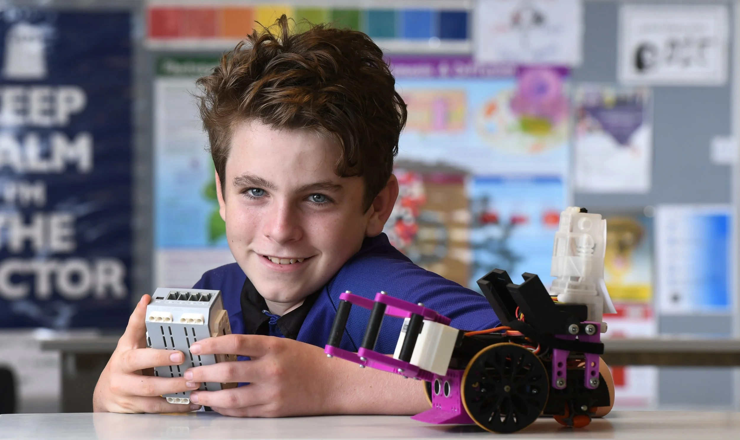 A young boy with brown curly hair smiling at the camera, holding a small electronic device, with a robot constructed from purple and black parts on the table in front of him, in a classroom with educational posters in the background.