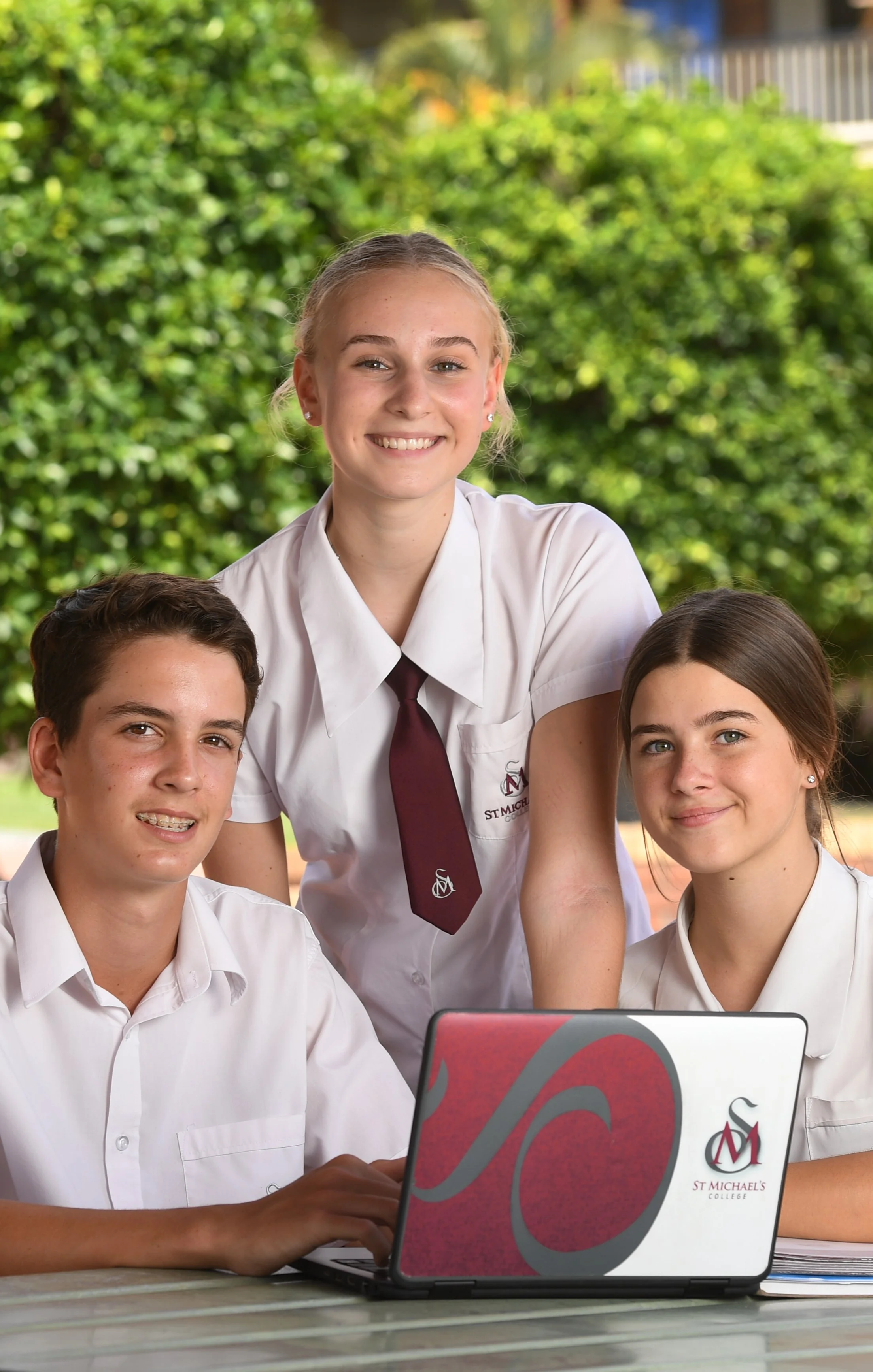 Three students in school uniforms, two boys and one girl, gathered around a laptop with the St. Michael's College logo, outdoors with green trees in the background.