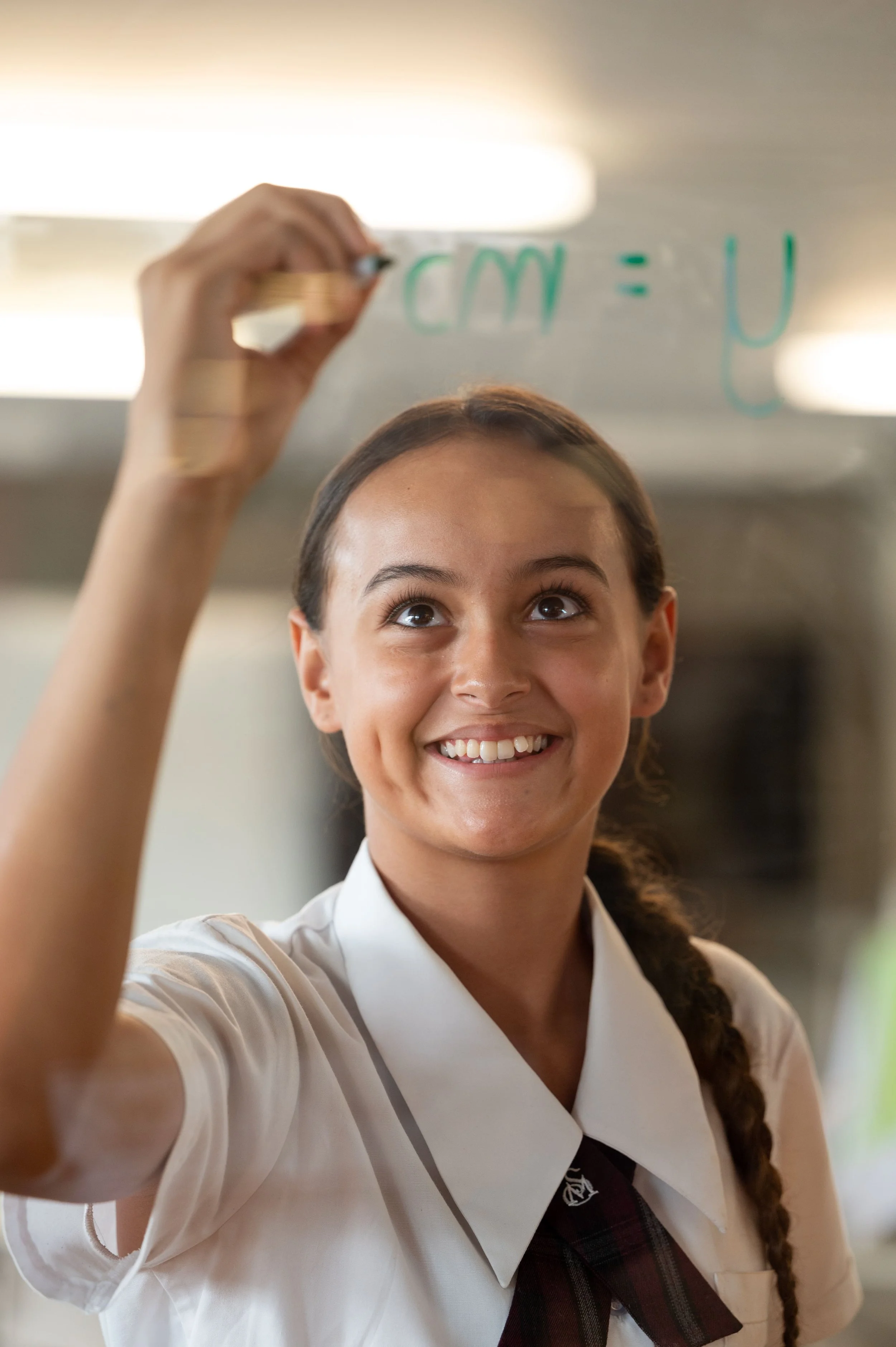A young girl in a school uniform smiling and writing on a transparent surface with a green marker, creating a math equation.