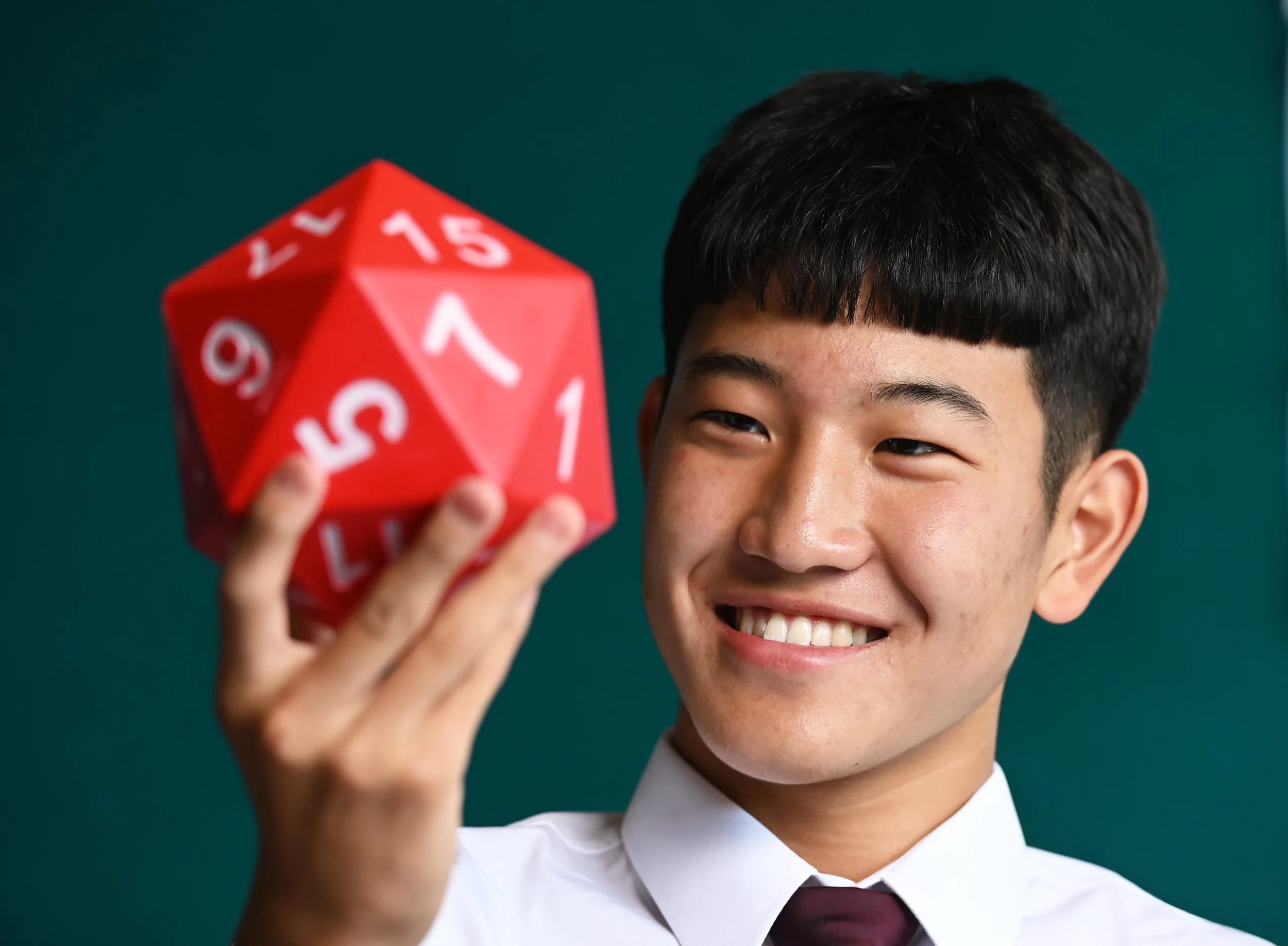 Young boy smiling and holding a red 20-sided die with white numbers against a green background.