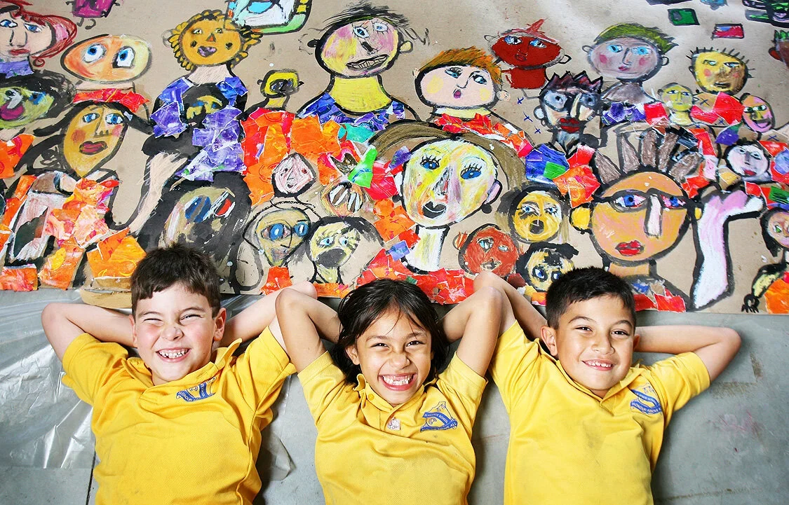 Three children in yellow school uniforms are lying on the floor with their hands behind their heads, smiling happily in front of a large colorful mural of abstract and expressive children's drawings. School and Education Marketing Photography