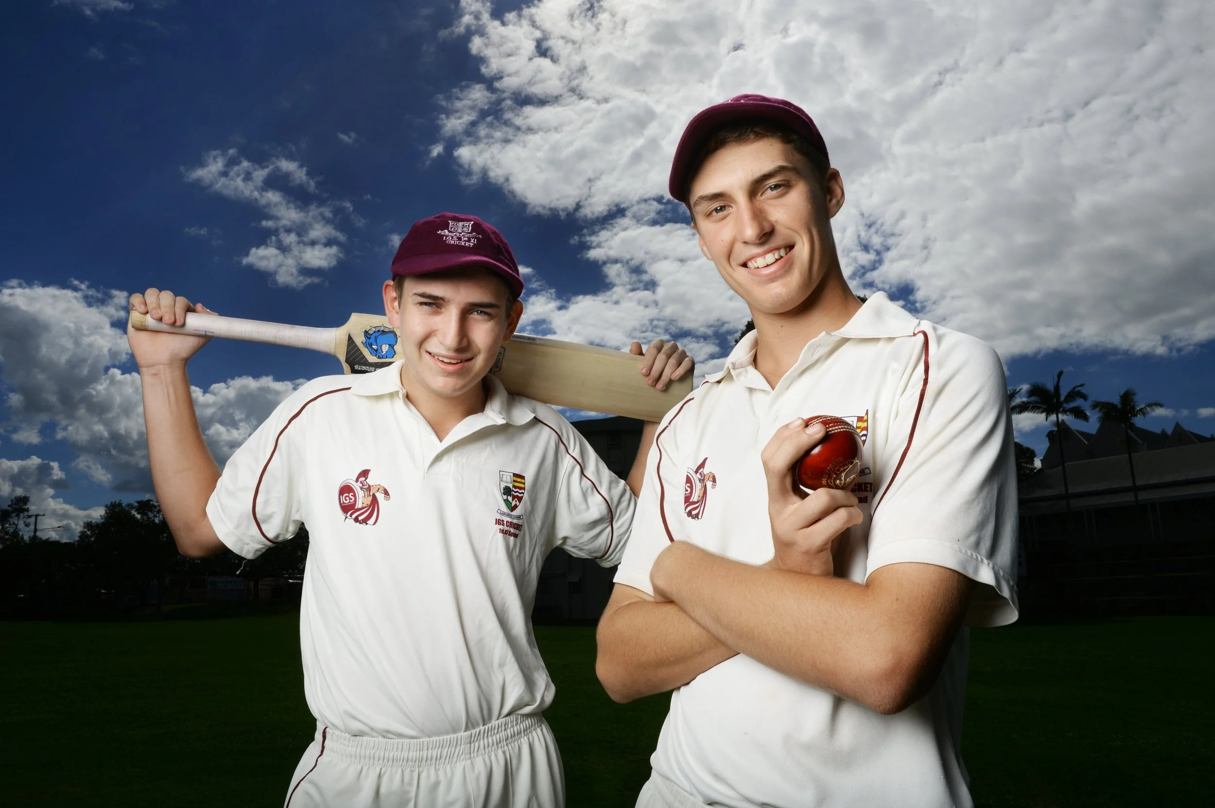 Two young male cricket players in white uniforms and maroon caps standing outdoors on a cricket field, one holding a cricket bat over his shoulder and the other holding a cricket ball, with a partly cloudy sky and trees in the background.