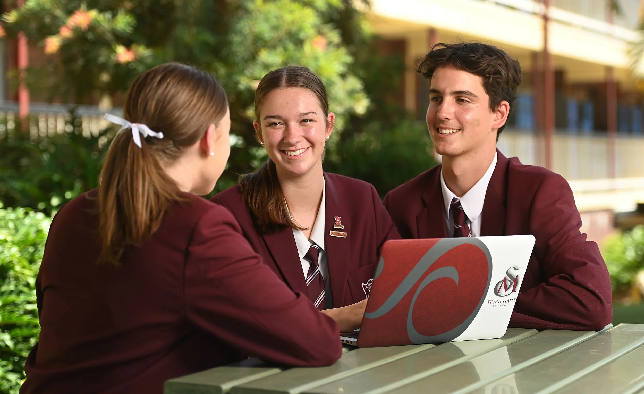 Three students in maroon blazers sitting at a table outdoors, talking and smiling, with a laptop displaying the St. Michael's College logo. School and Education Marketing Photography