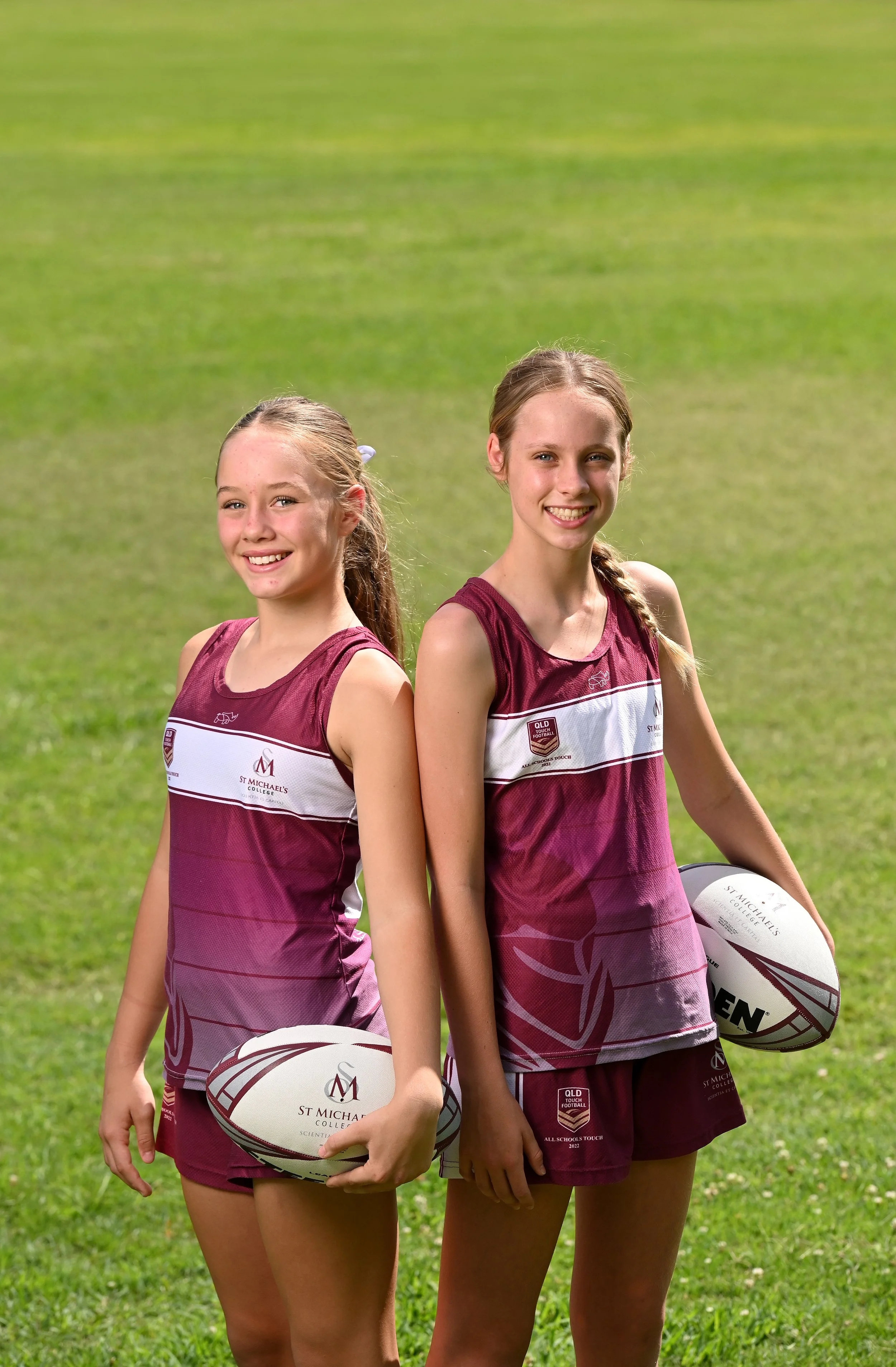 Two young girls in maroon and white rugby uniforms standing on a grassy field, each holding a rugby ball and smiling at the camera.