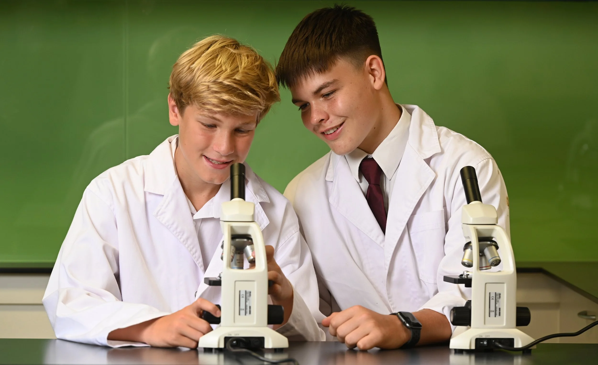Two boys in white lab coats looking into microscopes in a science classroom.