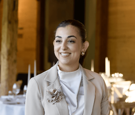 Eine lächelnde Frau in einem hellen Blazer und weißem Oberteil, im Hintergrund ein elegantes Restaurant