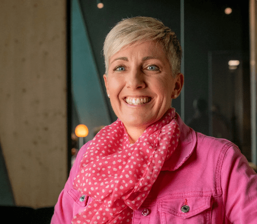 Eine smiling Frau mit kurzen, grauen Haaren trägt eine pinke Jacke und ein pinkes Halstuch in einem modernen, dunklen Raum.