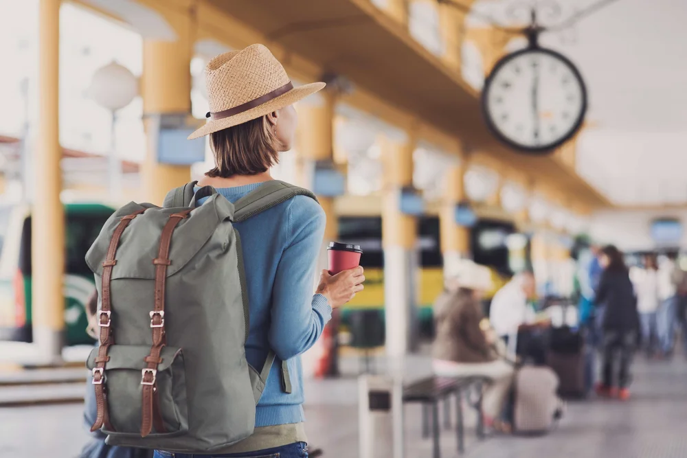 Une femme avec un sac à dos et un chapeau de paille regarde l’horloge d’une gare, un café à la main
