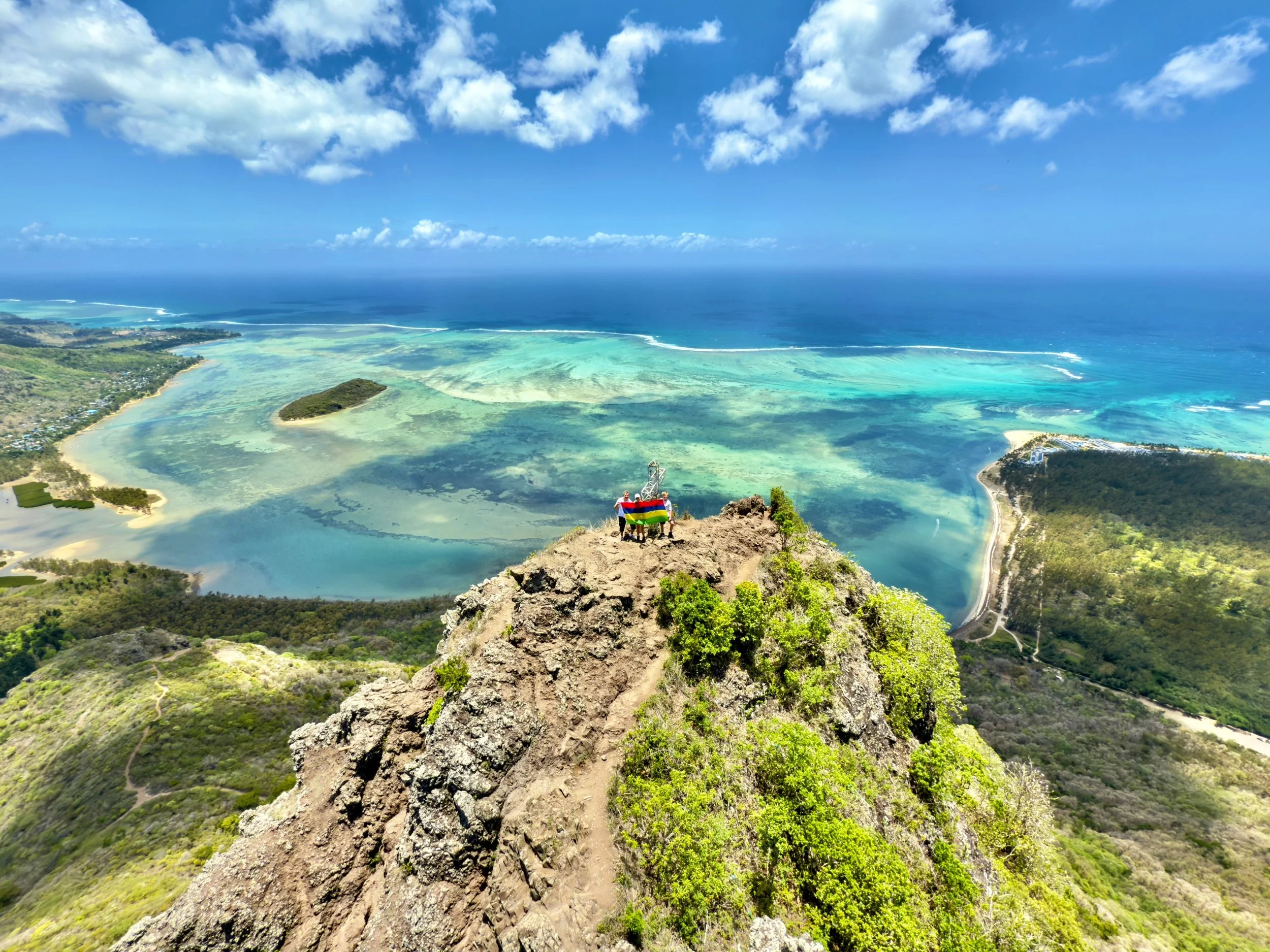 Scenic view from Le Morne Brabant Mountain overlooking turquoise ocean and beach, with hikers on the summit.