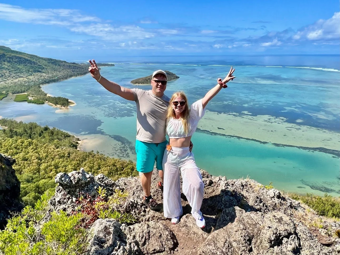 A couple posing on a rocky viewpoint overlooking a turquoise lagoon and lush landscape with mountains in the background.