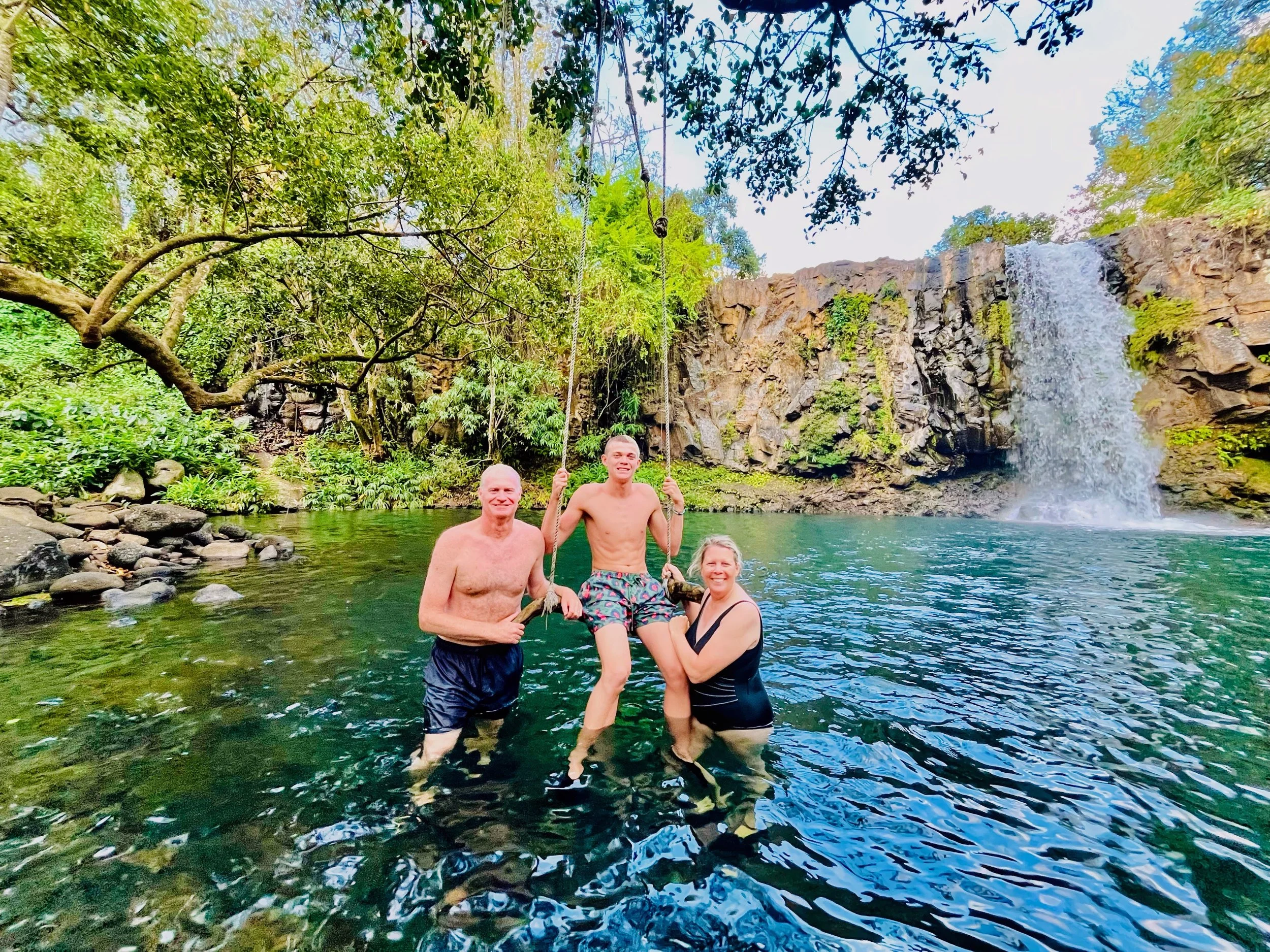 Group of people standing in a natural pool with a waterfall and lush greenery in the background.