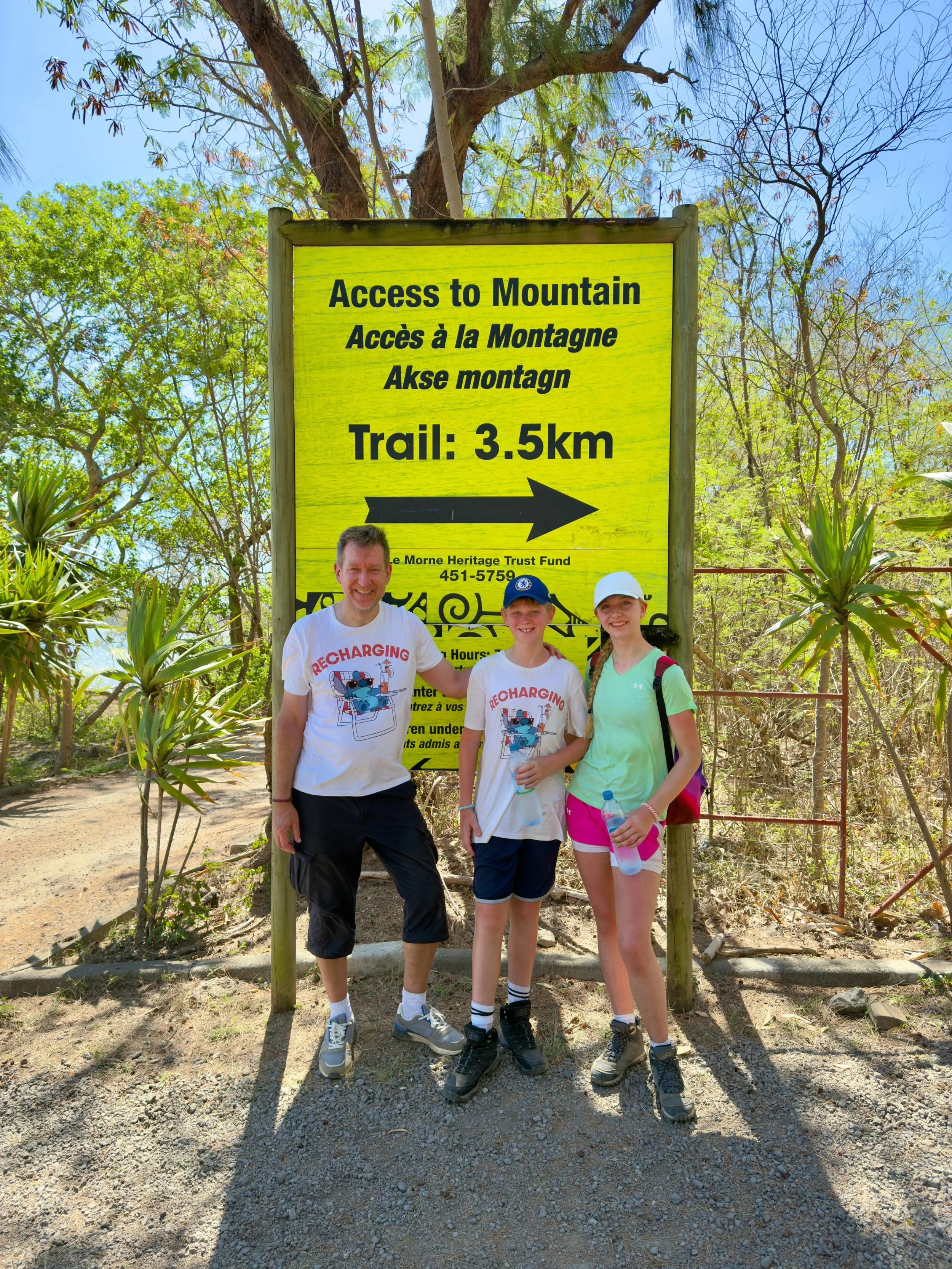 Three people posing in front of a yellow sign in a natural setting. The sign reads 'Access to Mountain' and indicates a trail of 3.5 km. Trees and plants are visible in the background.