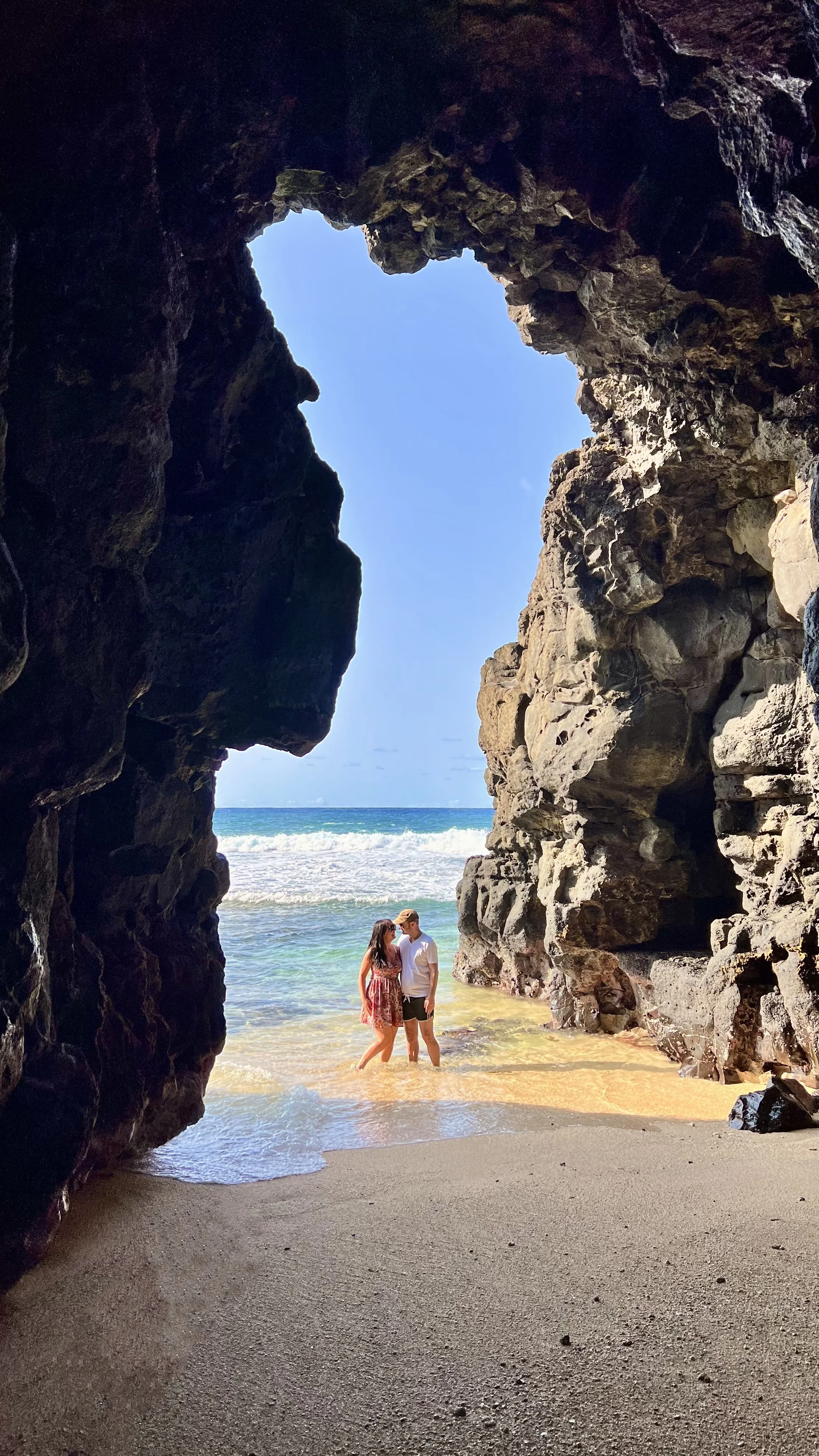 Couple standing on a sandy beach inside a rocky sea cave with ocean view.