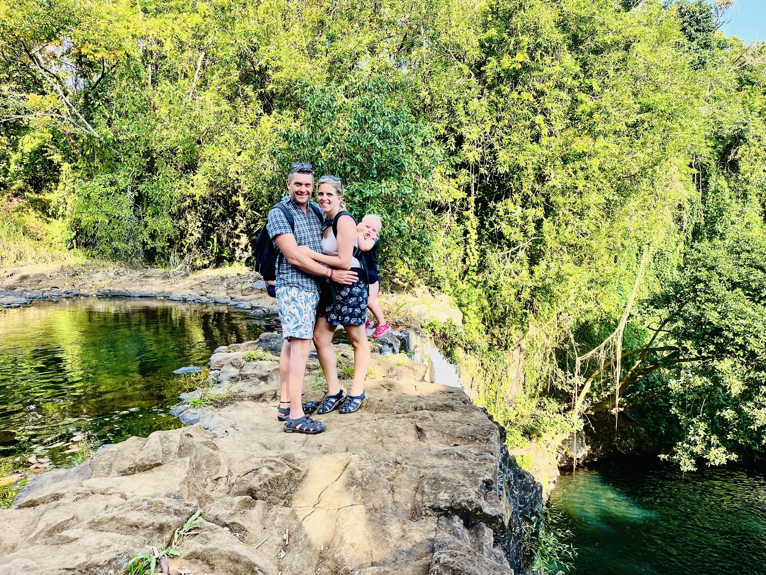 Three people standing on rocky pathway near a pond with lush green trees in the background.
