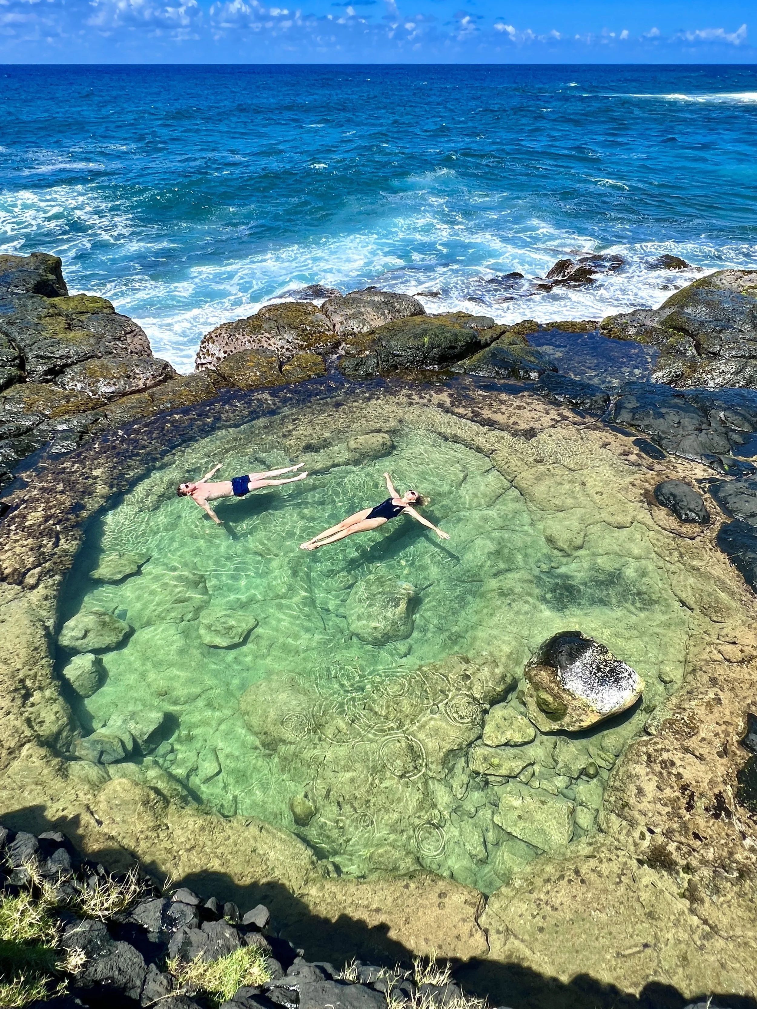 Two people floating in a natural tide pool surrounded by rocks, with ocean waves visible in the background.