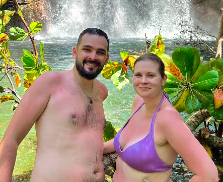Two people standing in front of a waterfall surrounded by lush greenery.