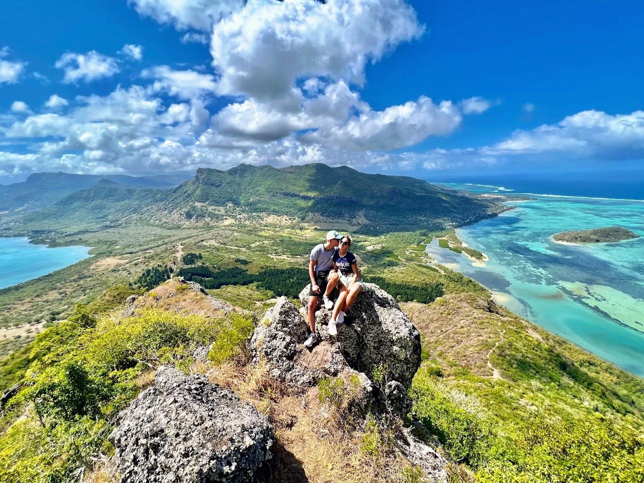 Two people sitting on a rocky cliff overlooking a scenic landscape with lush green hills, turquoise ocean, and blue sky with clouds in the background.