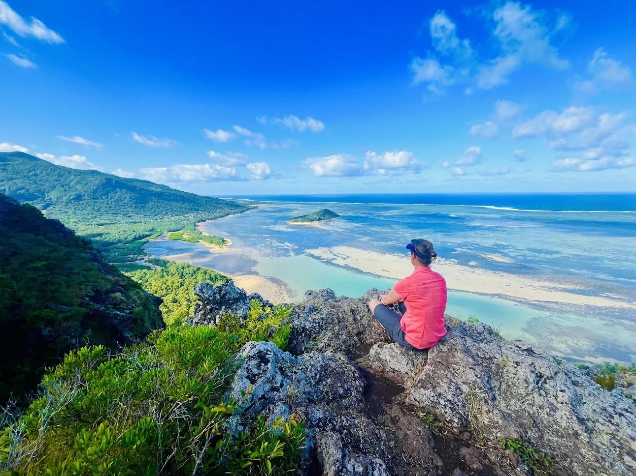 Person sitting on a cliff overlooking a coastal landscape with mountains, sea, and cloudy sky.