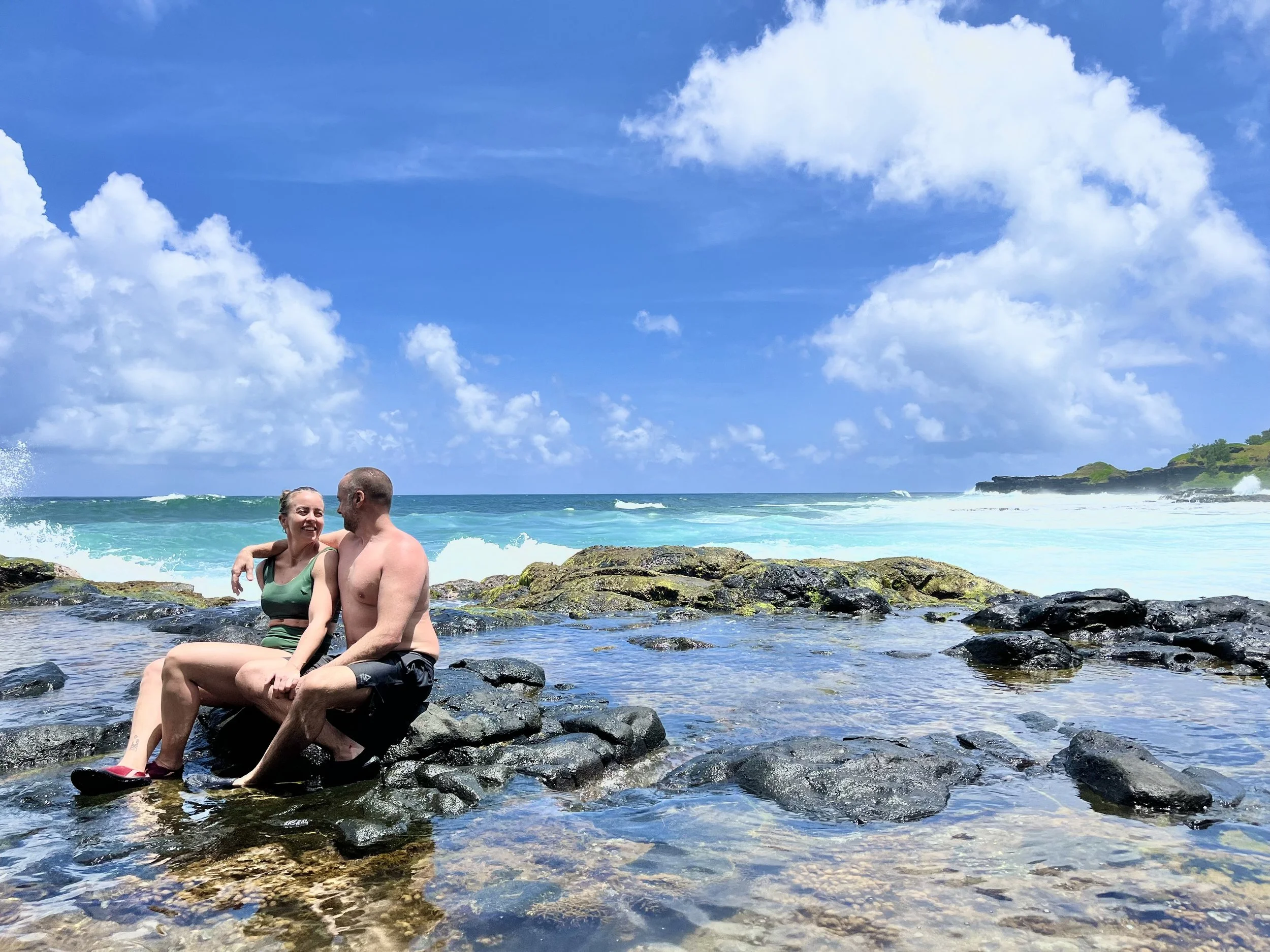 Couple sitting on rocks by the ocean with blue sky and clouds