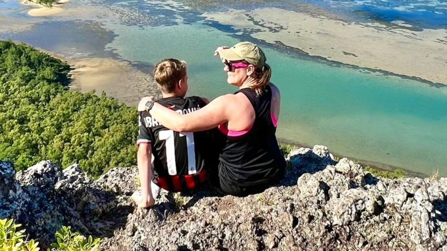 Mother and son sitting on rocky ledge overlooking a scenic coastal view with green foliage and turquoise waters.