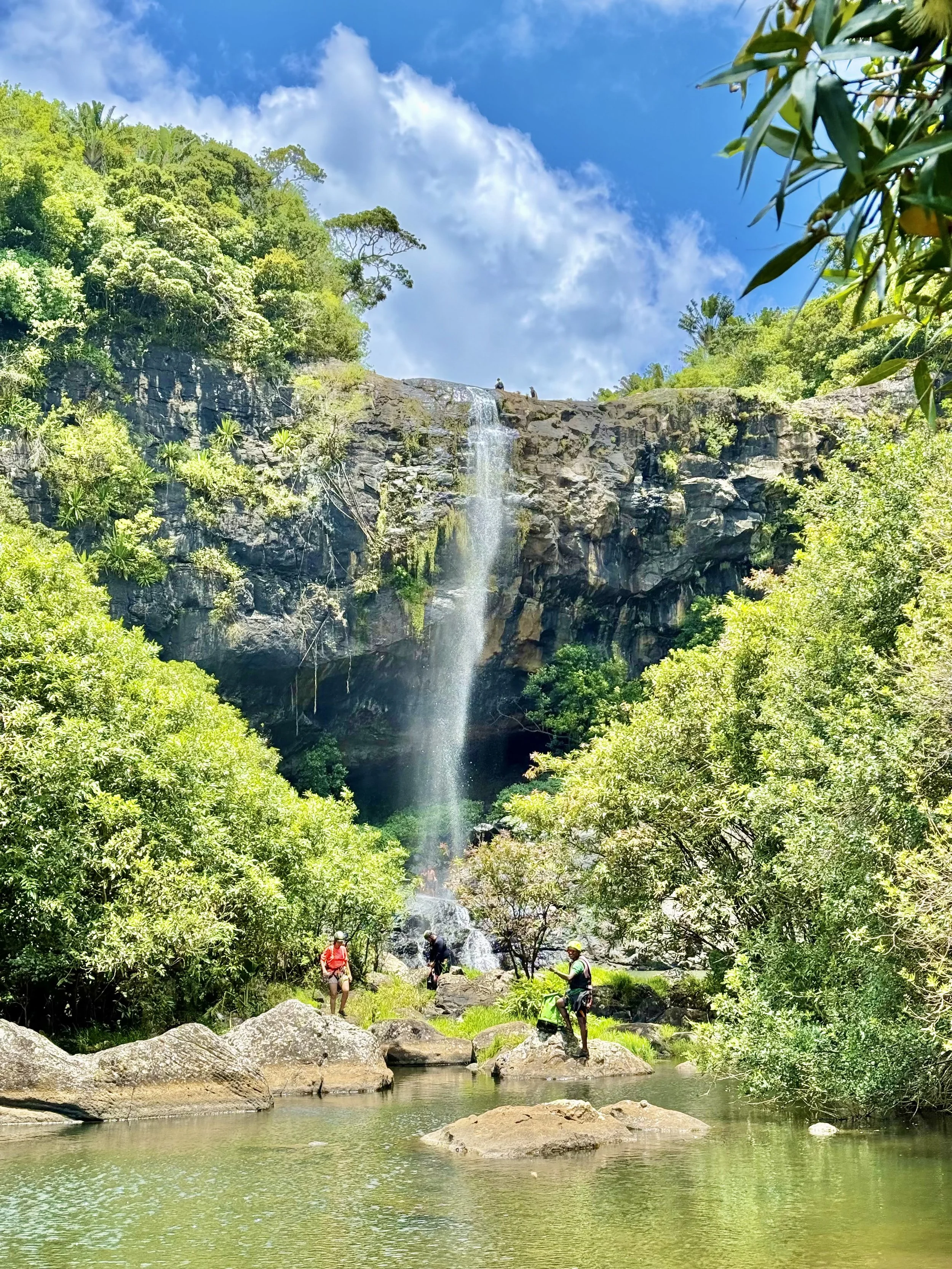 Scenic waterfall with hikers nearby, surrounded by lush greenery and rocky terrain, under a bright blue sky.