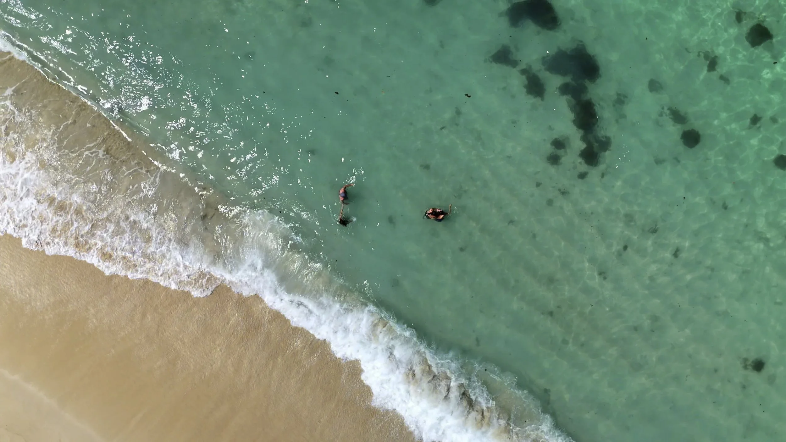 Aerial view of two people swimming in clear turquoise ocean near a sandy beach.
