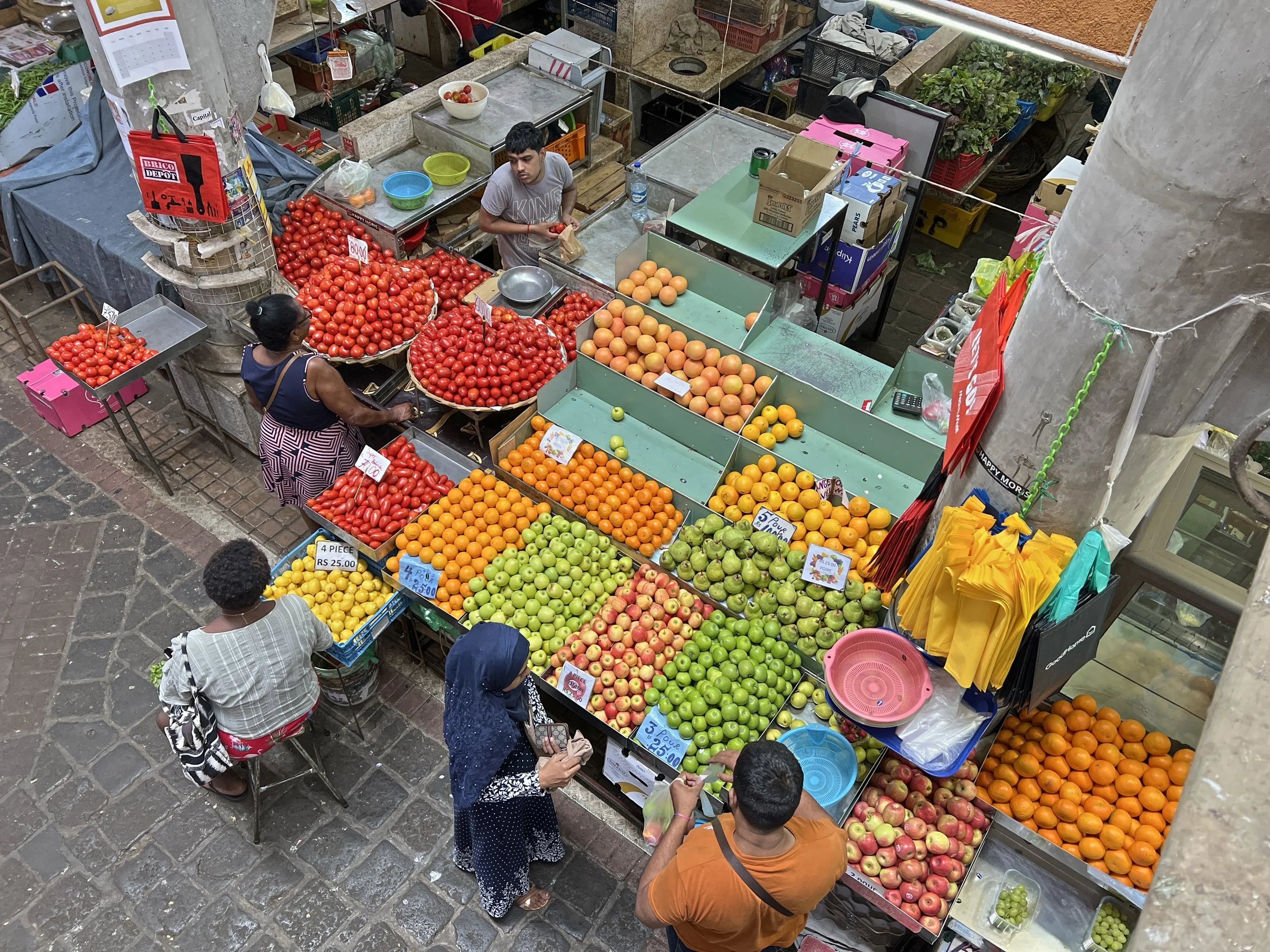 Aerial view of a bustling fruit market featuring piles of tomatoes, oranges, green apples, and other fruits with vendors assisting customers.