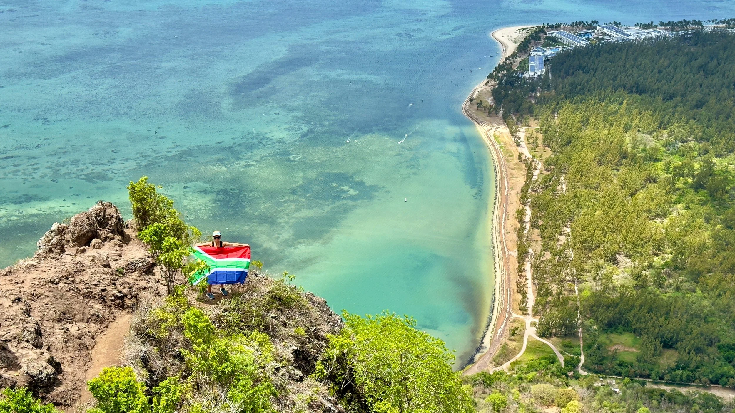Scenic view from Le Morne Brabant viewpoint with hiker holding Mauritius flag, overlooking turquoise lagoon, coastline, and greenery.