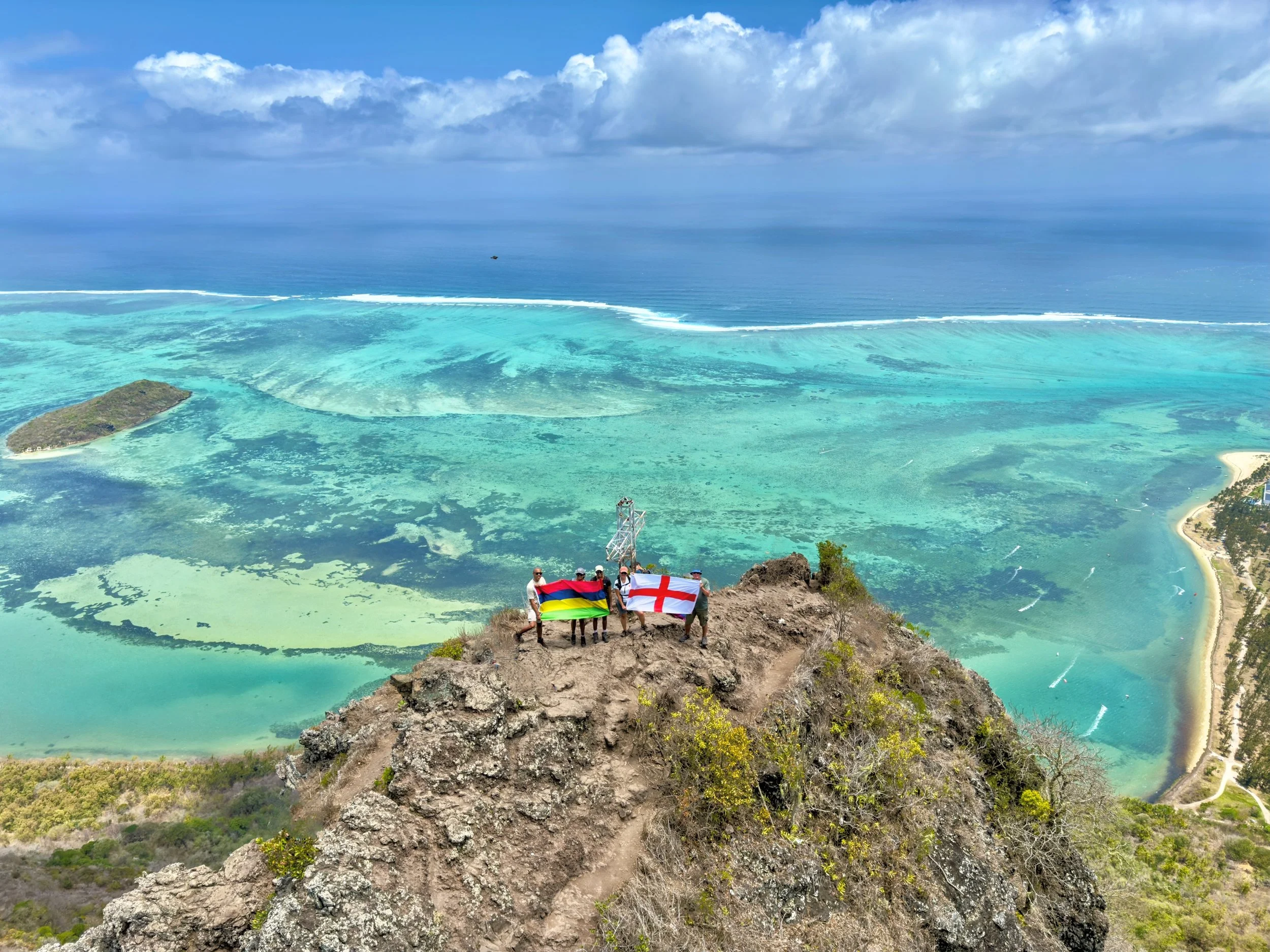 Scenic view from Lion Mountain in Mauritius overlooking turquoise ocean waters, small island, and coastline; hikers holding flags.