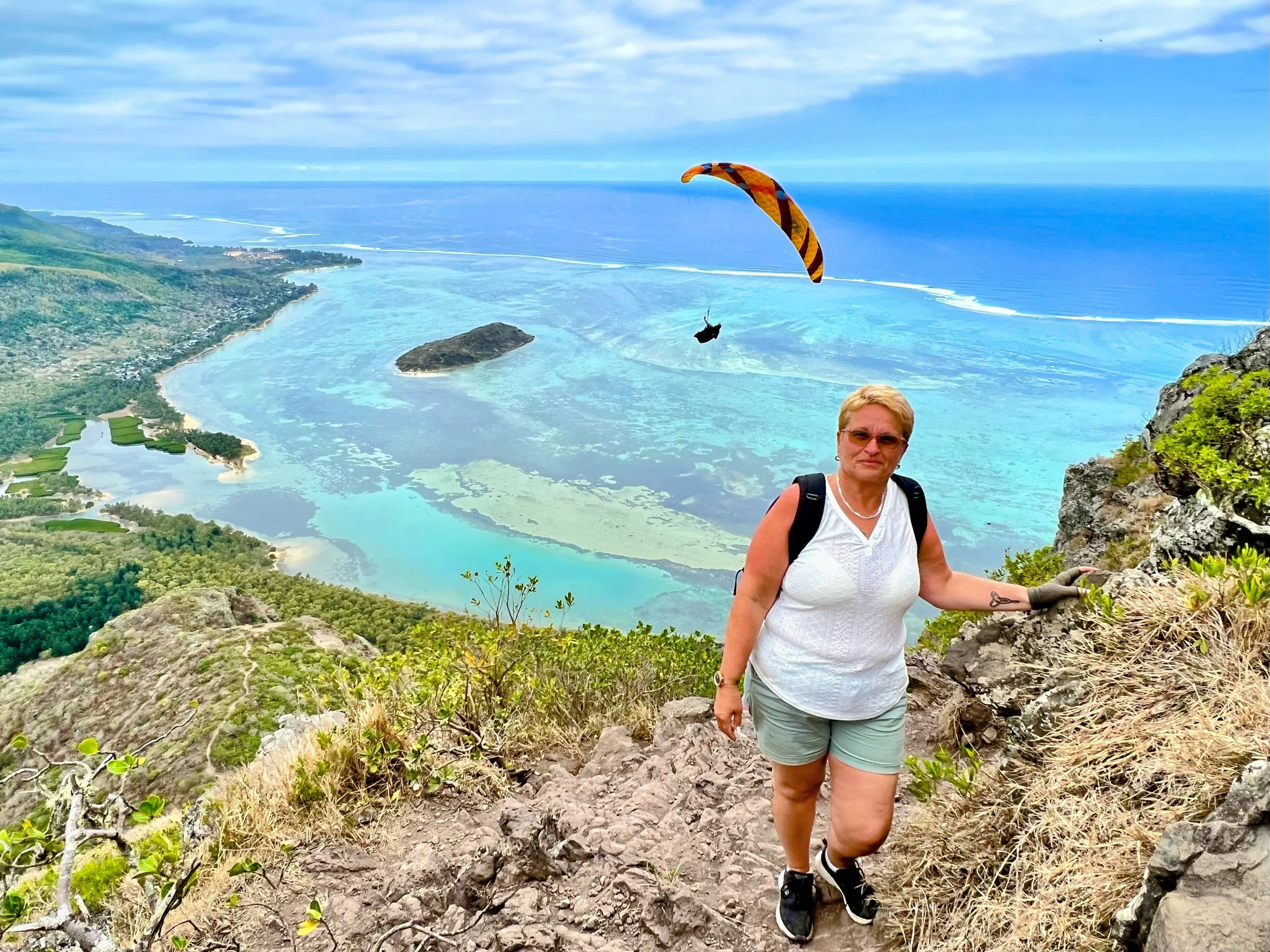 Woman hiking on rocky trail with tropical coastline and paraglider in background.