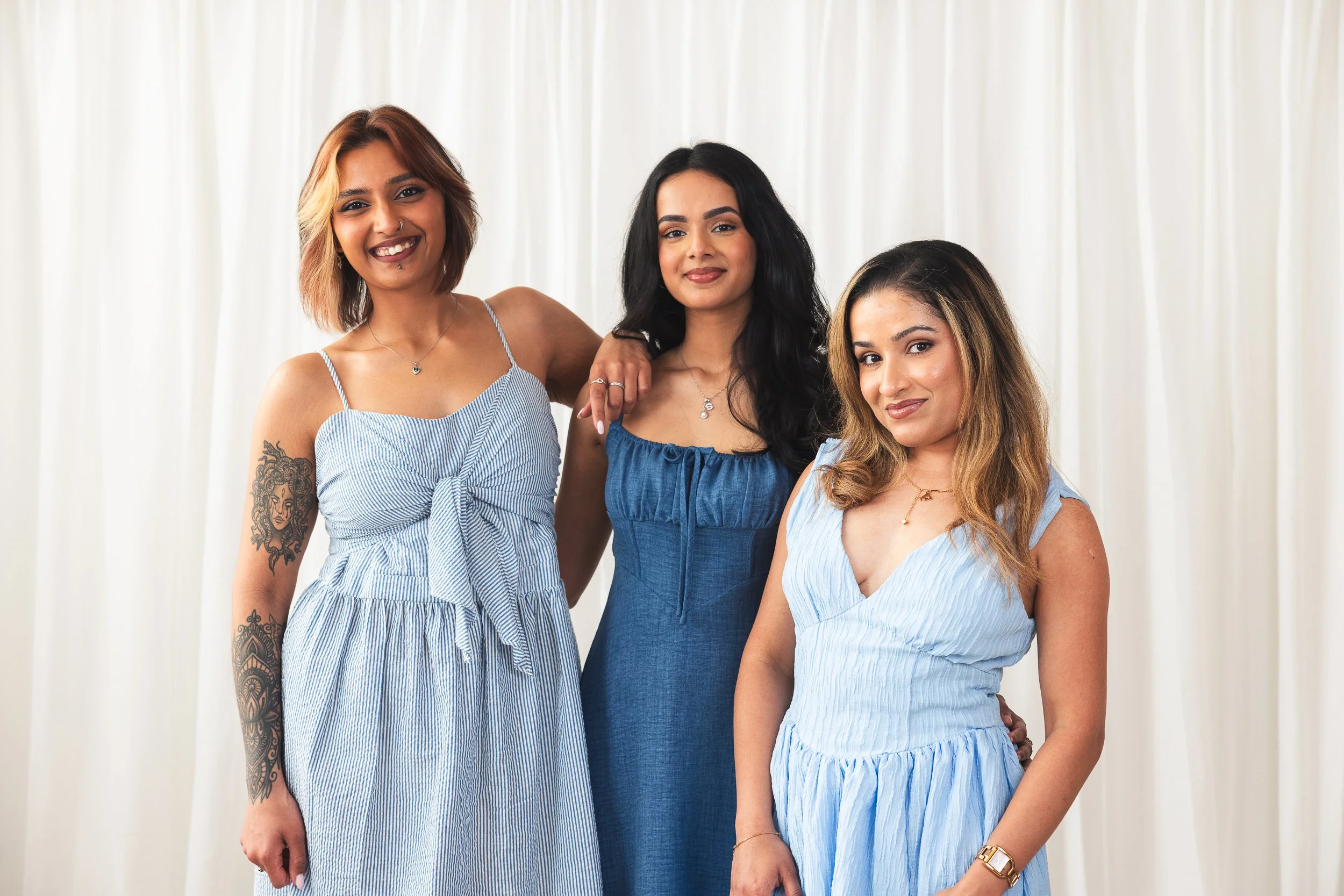 Three women dressed in summer dresses standing together against a white curtain backdrop, smiling at the camera.